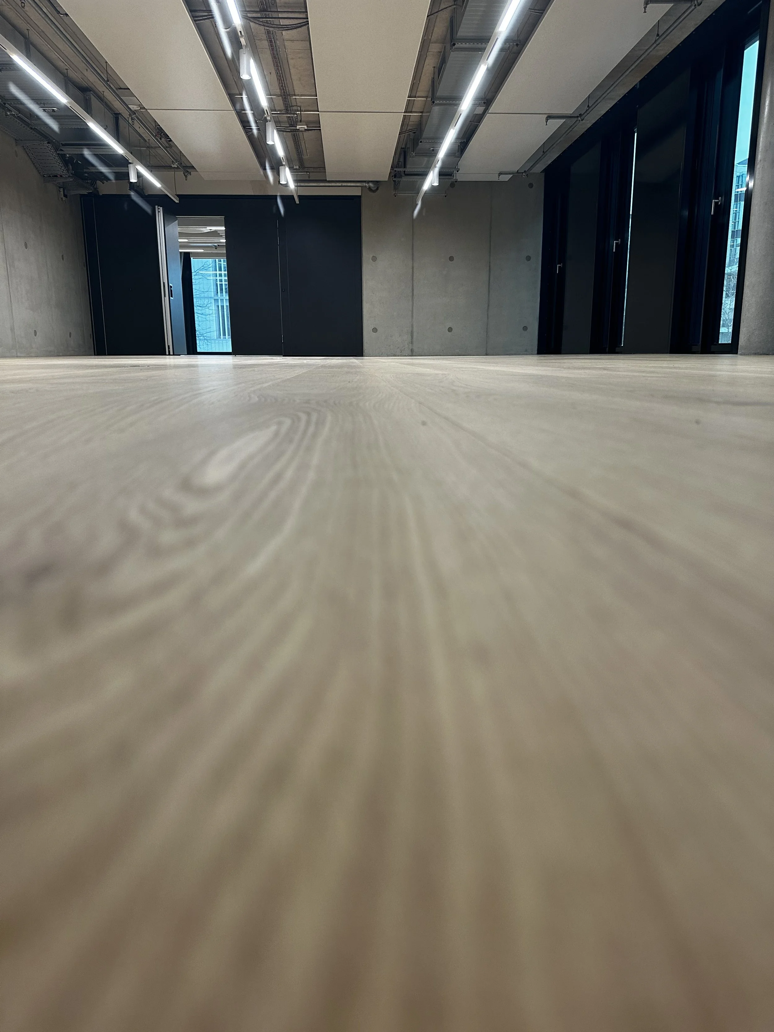 Innenraum eines modernen Büros mit Holztisch, Betonwänden, schwarzen Türen, großen Fenstern und Deckenbeleuchtung.