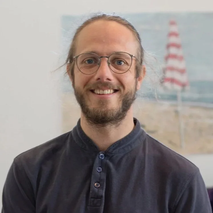 Lächelnder Mann mit Brille und dunklem Hemd vor einem Strandbild mit Sonnenschirm.