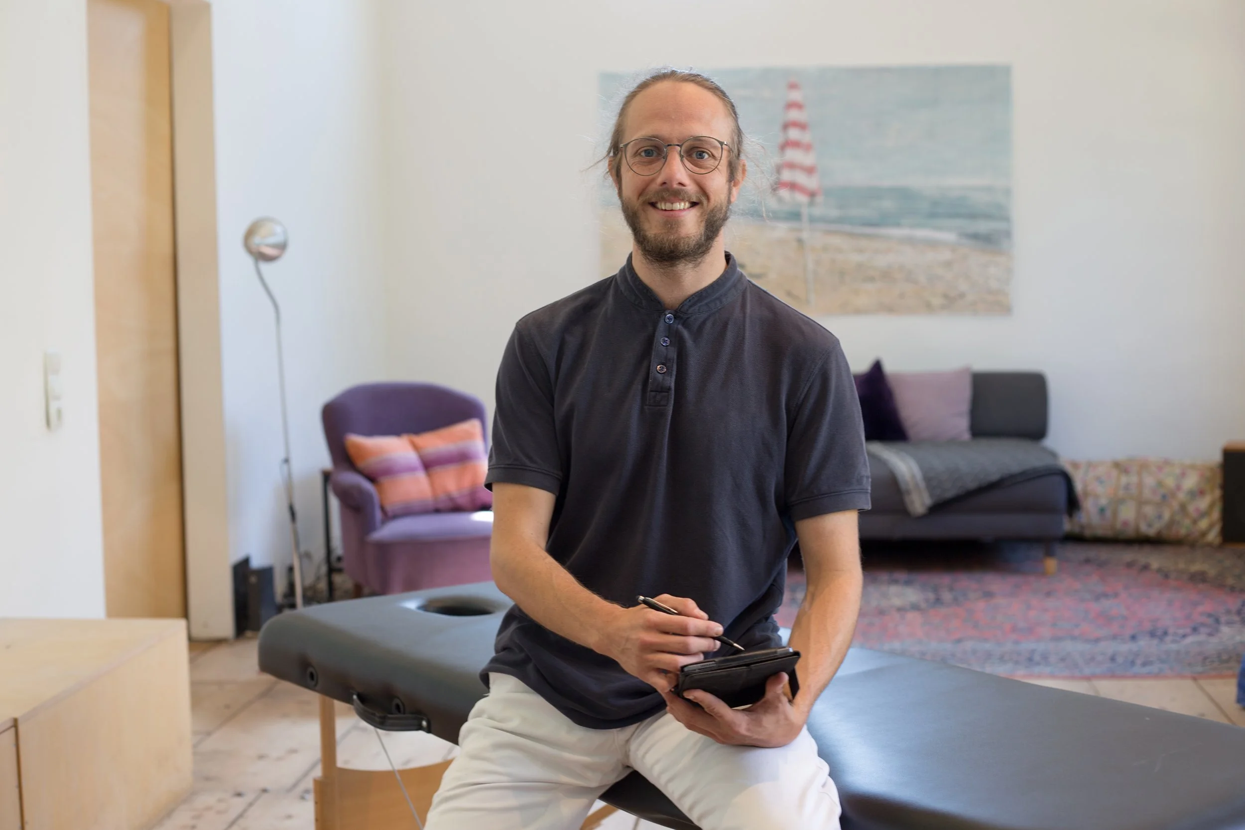 Ein Mann mit Brille und Bart sitzt auf einer Behandlungsliege in einem Raum, der wie eine Praxis aussieht, und hält einen digitalen Notizblock und einen Stift, im Hintergrund ein Sofa und ein Bild vom Strand mit einem roten und weißen Sonnenschirm.