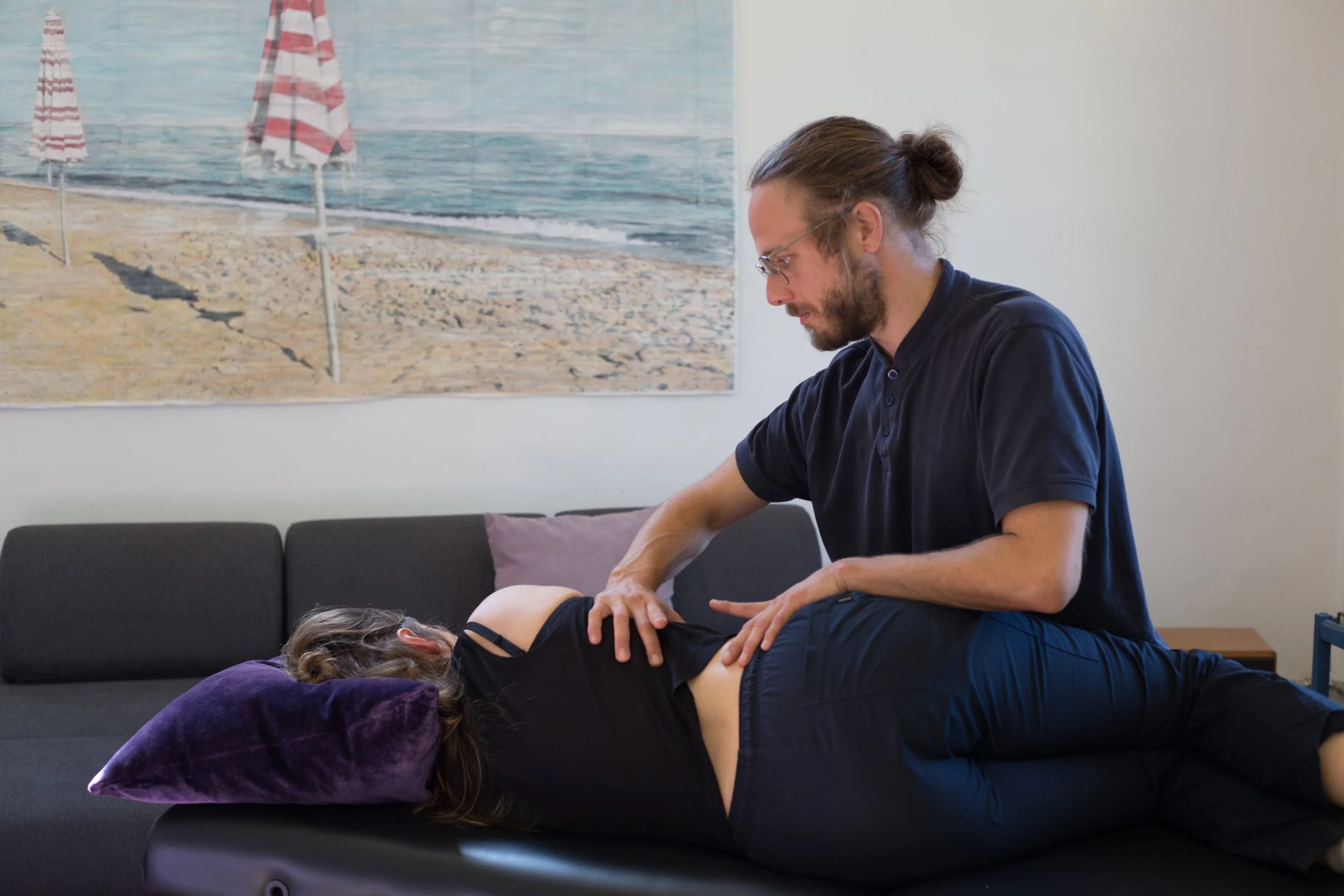 Ein Mann führt eine Chiropraktik-Behandlung an einer liegenden Frau durch, die auf einer schwarzen Behandlungsliege liegt. Im Hintergrund ist ein Wandbild eines Strandes mit Sonnenliegen und Sonnenschirmen.