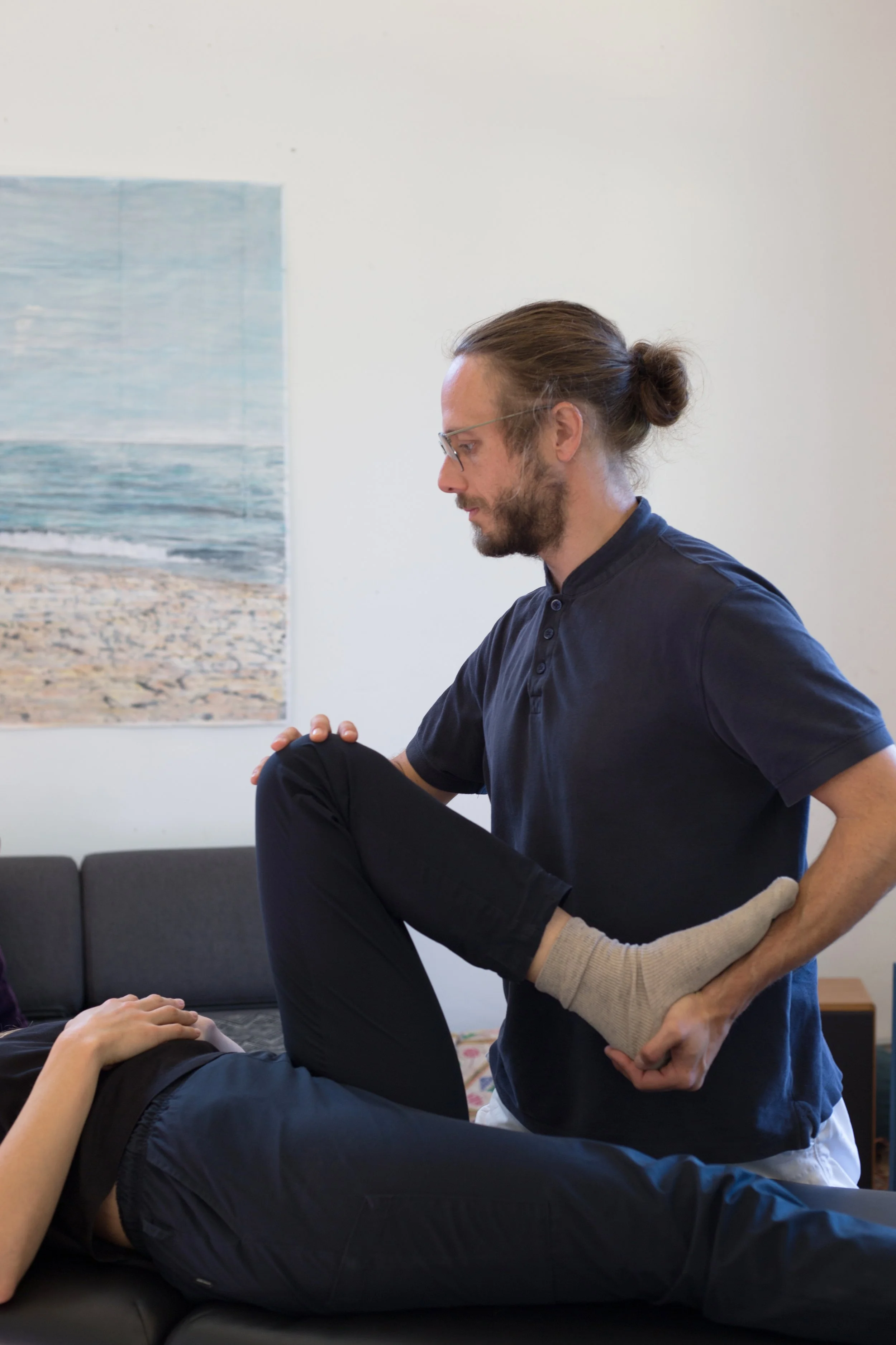 Ein Mann mit Brille führt eine physische Therapie an einem sitzenden Patienten durch, der auf einer Behandlungsliege liegt.