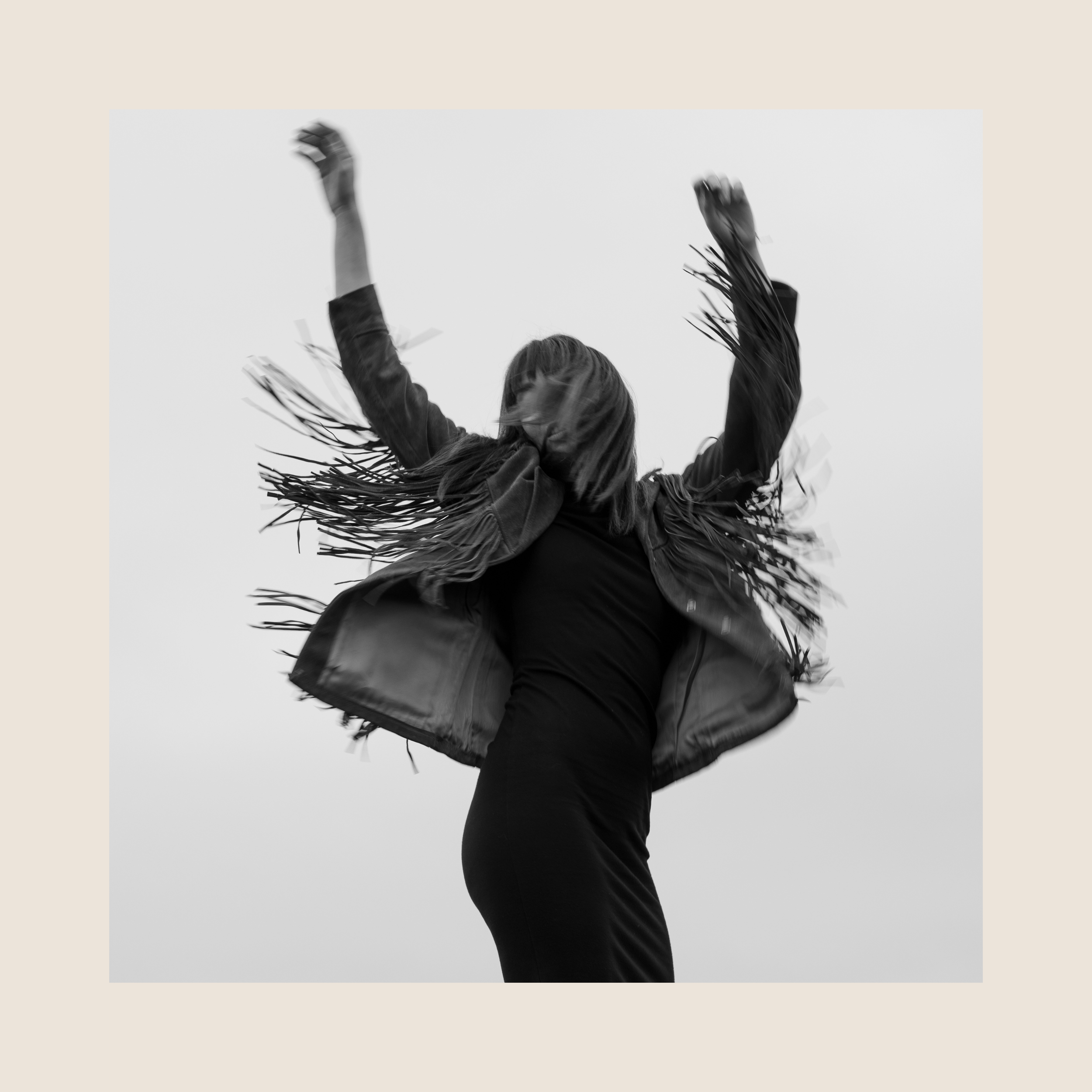 Une femme en mouvement avec les bras levés, portant une veste à franges, vue de dos, en noir et blanc.