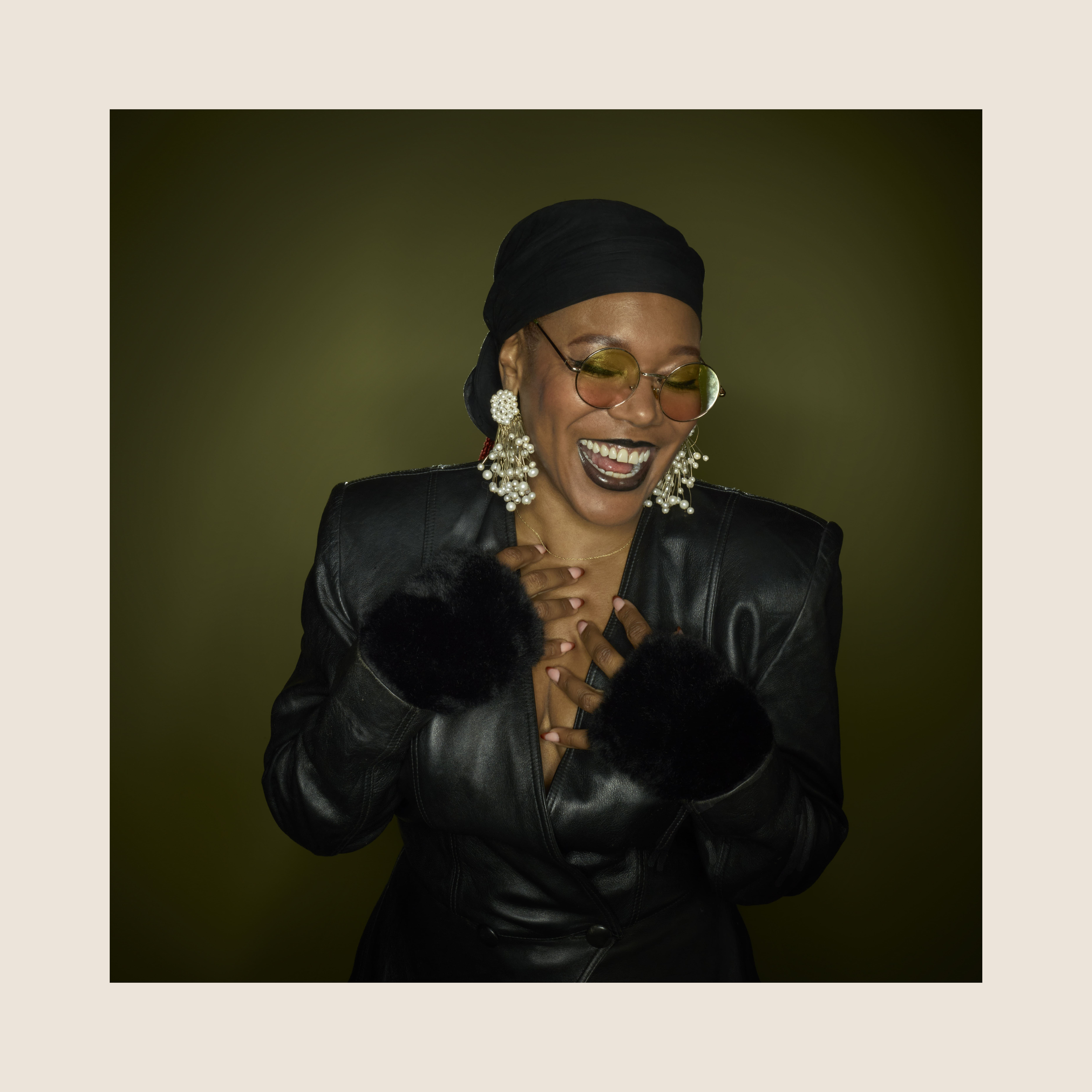 Femme souriante portant des lunettes de soleil rondes, des boucles d'oreilles en perles, une veste en cuir avec des poignets en fourrure noire, et un bandeau noir, posant avec une main sur la poitrine contre un fond vert foncé.