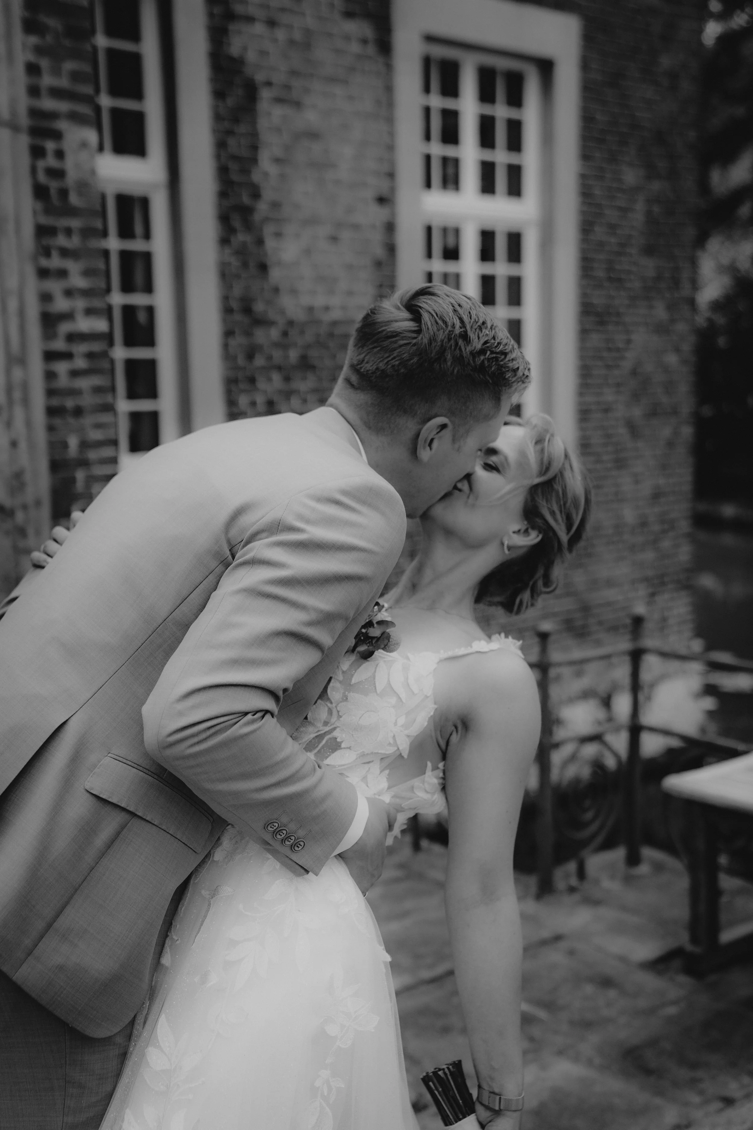 Ein Brautpaar küsst sich an einer Häuserwand im Freien, Schwarz-Weiß-Foto, Hochzeitskleidung, romantische Szene.