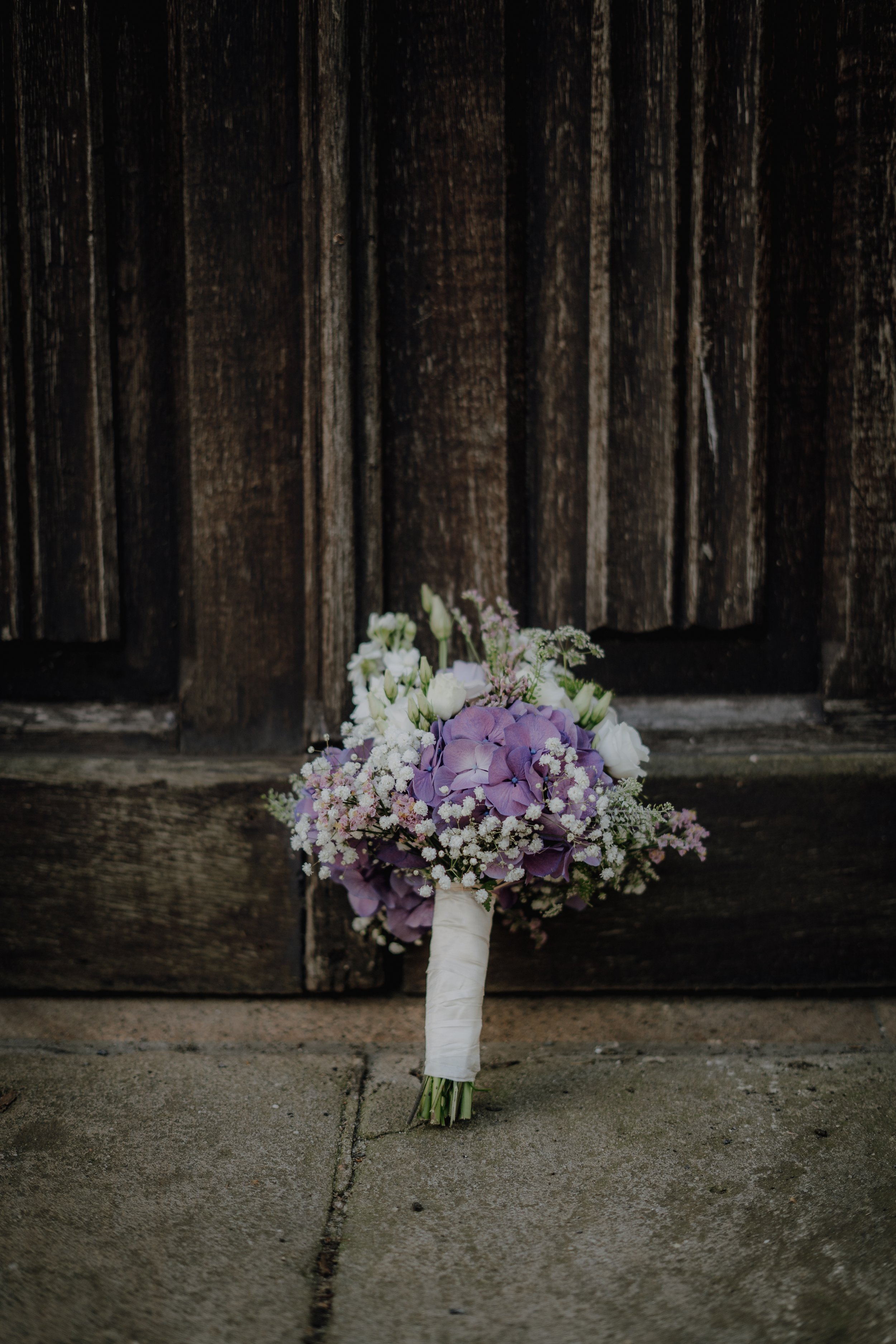 Blumenstrauß mit lilafarbenen und weißen Blumen, vor einem dunklen Holzschrank auf dem Boden liegend.
