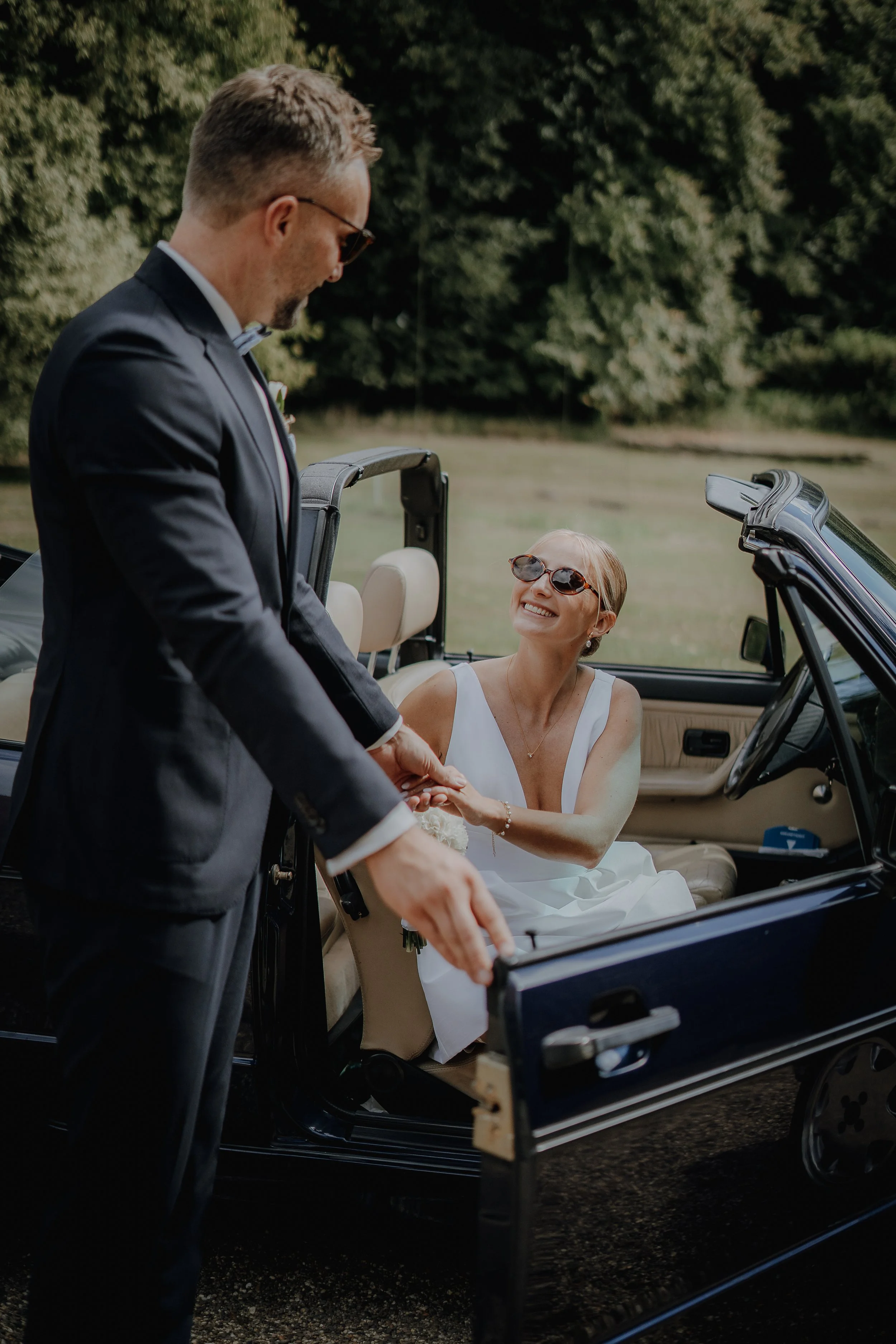 Ein Mann im Anzug hilft einer lachenden Frau in einem weißen Kleid beim Einsteigen in ein Cabriolet, umgeben von Natur.