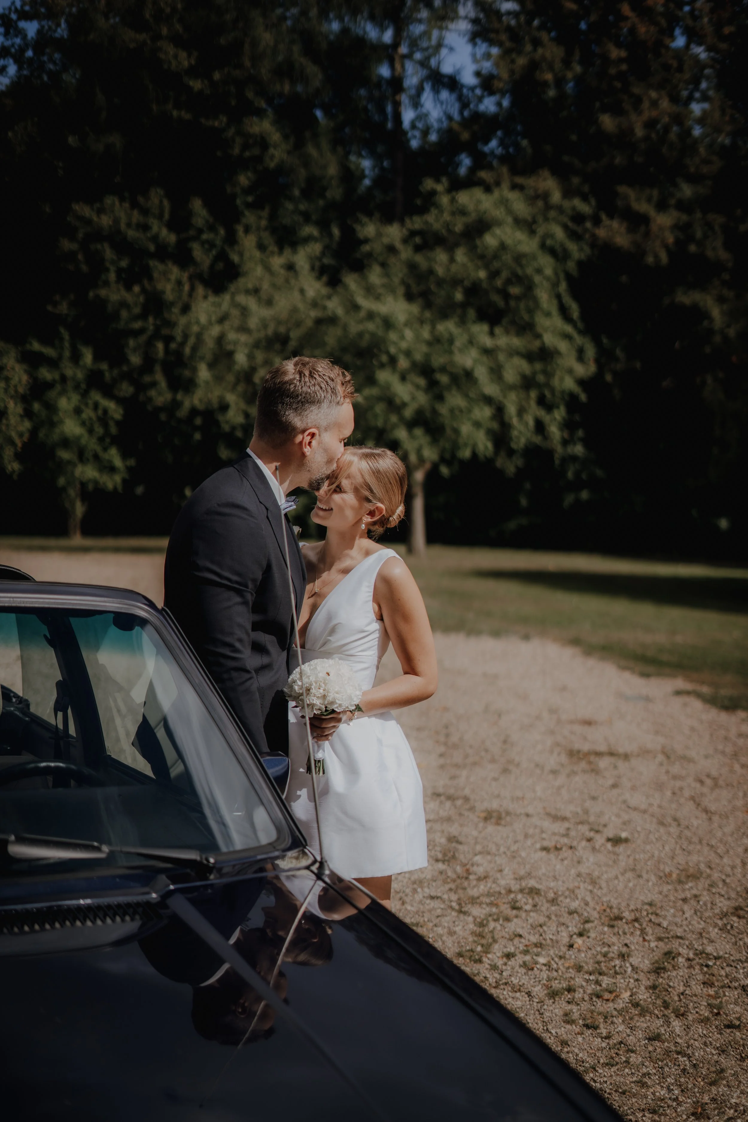 Ein Brautpaar steht neben einem schwarzen Auto im Freien, lächelt sich an und küsst sich. Die Braut trägt ein weißes Kleid und hält einen Blumenstrauß, der Bräutigam trägt einen schwarzen Anzug. Im Hintergrund sind Bäume und eine Wiese zu sehen.