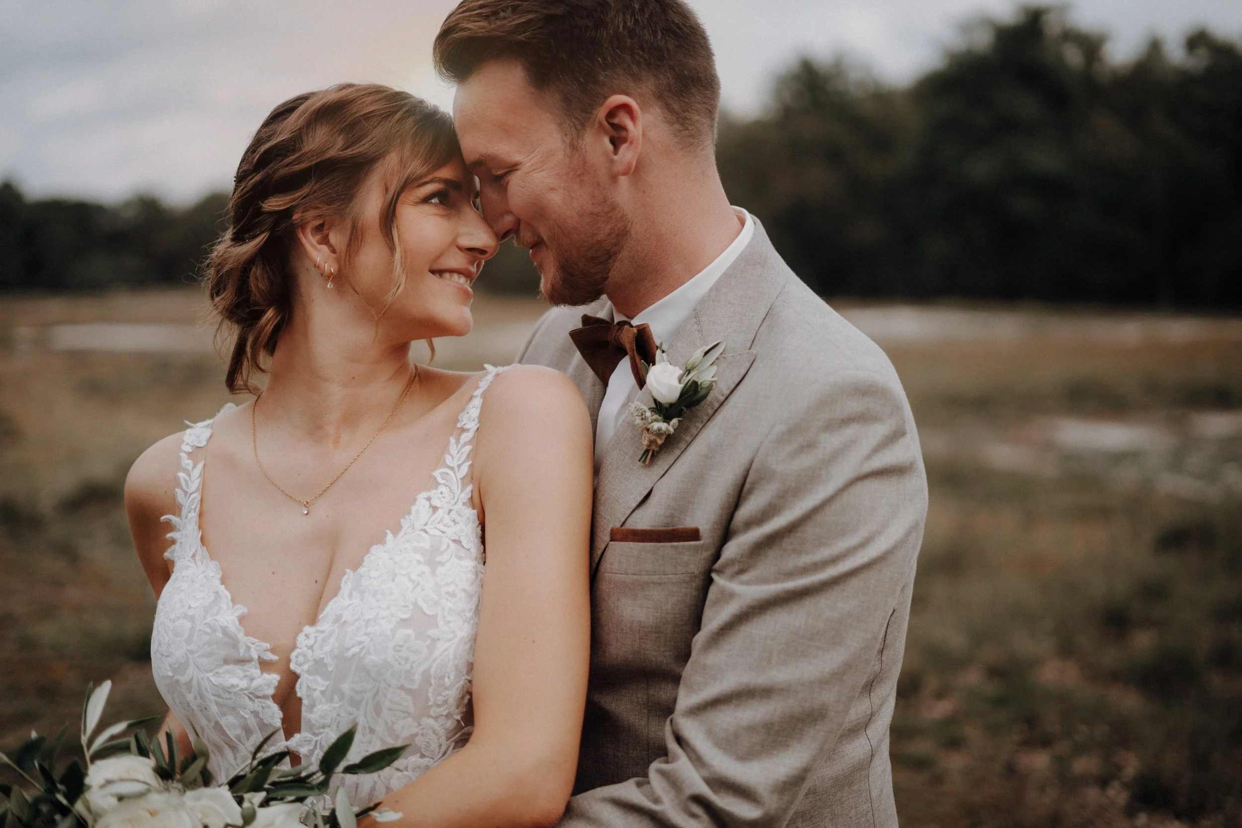 Ein glückliches Brautpaar, das sich an einem offenen Landschaftsweg bei Sonnenuntergang umarmt, mit einem sanften Lächeln, im Hochzeitskleid und Anzug.