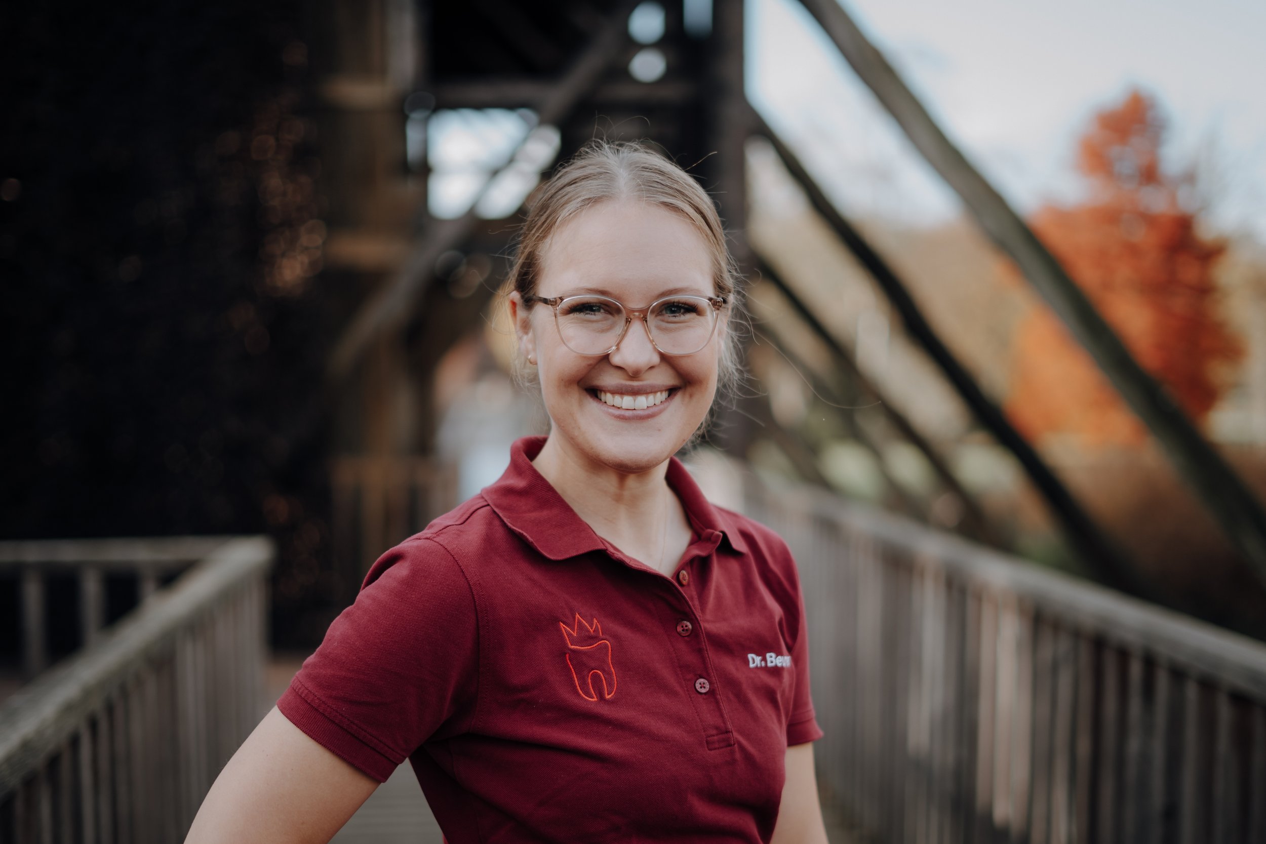 Eine lächelnde Frau mit Brille trägt ein rotes Polo mit einer Zahn- und Kronen-Logo und steht auf einer Brücke im Freien, im Hintergrund sind bunte Herbstbäume.