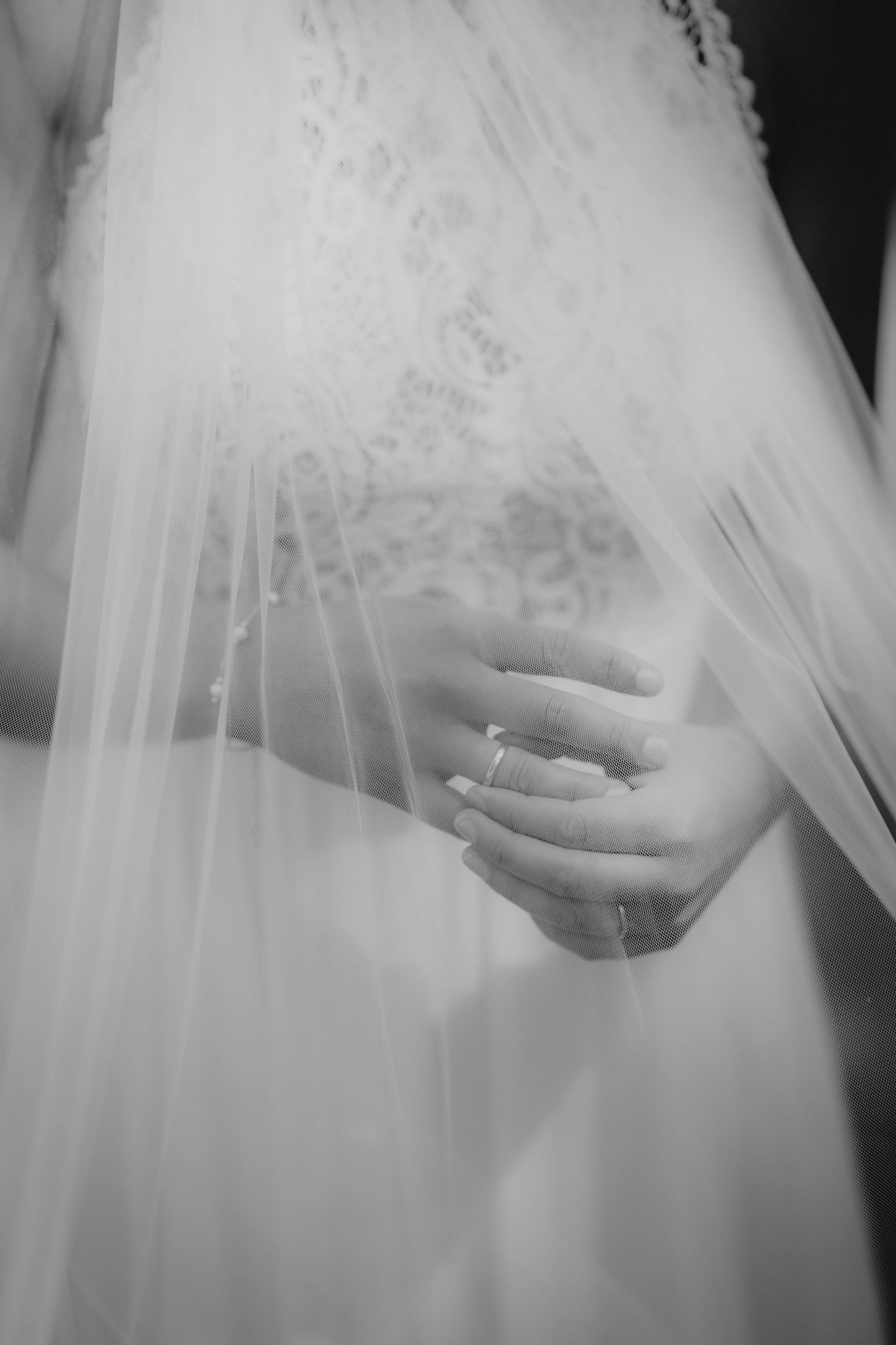 Schwarz-weiß-Foto einer Hand, die einen Ring trägt, unter einem Schleier auf einem Brautkleid mit Spitze.