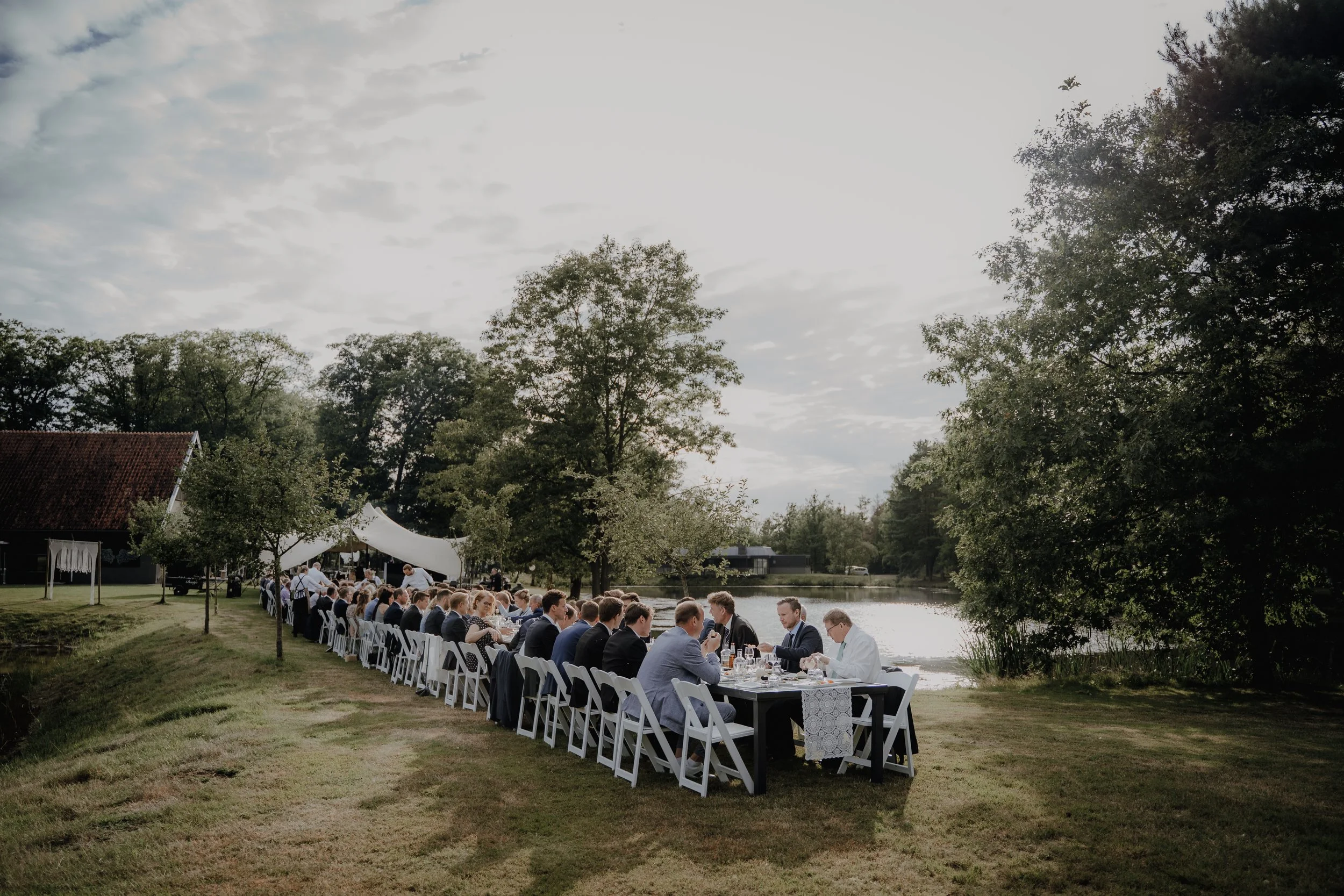 Gruppe von Menschen, die an einem langen Esstisch im Freien neben einem Fluss bei Sonnenuntergang speisen und feiern.