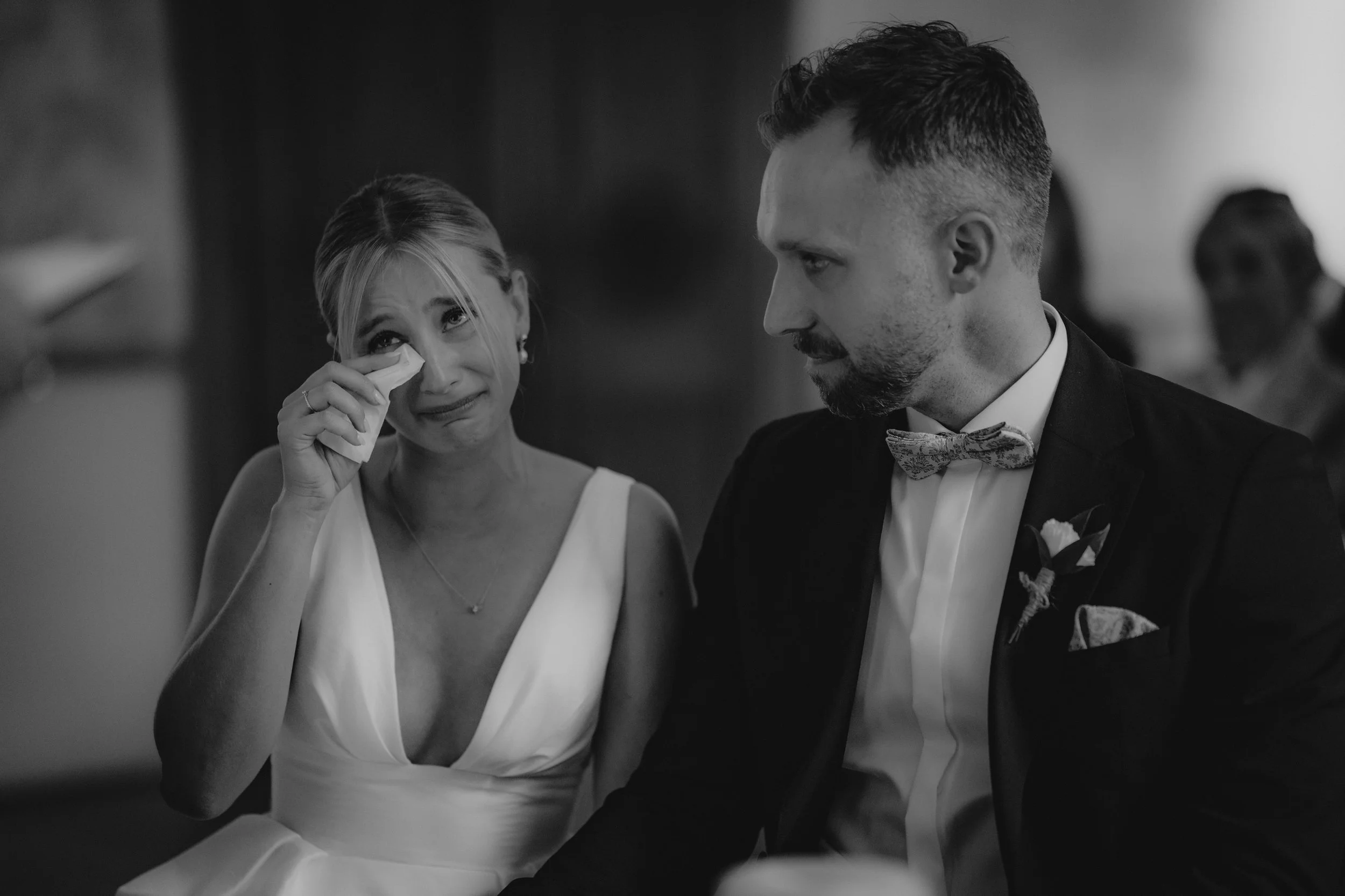 Eine traurige Frau in einem weißen Kleid weint, während sie an einer Hochzeit mit einem Mann in einem Anzug sitzt, der sie tröstet. Das Bild ist in Schwarz-Weiß.