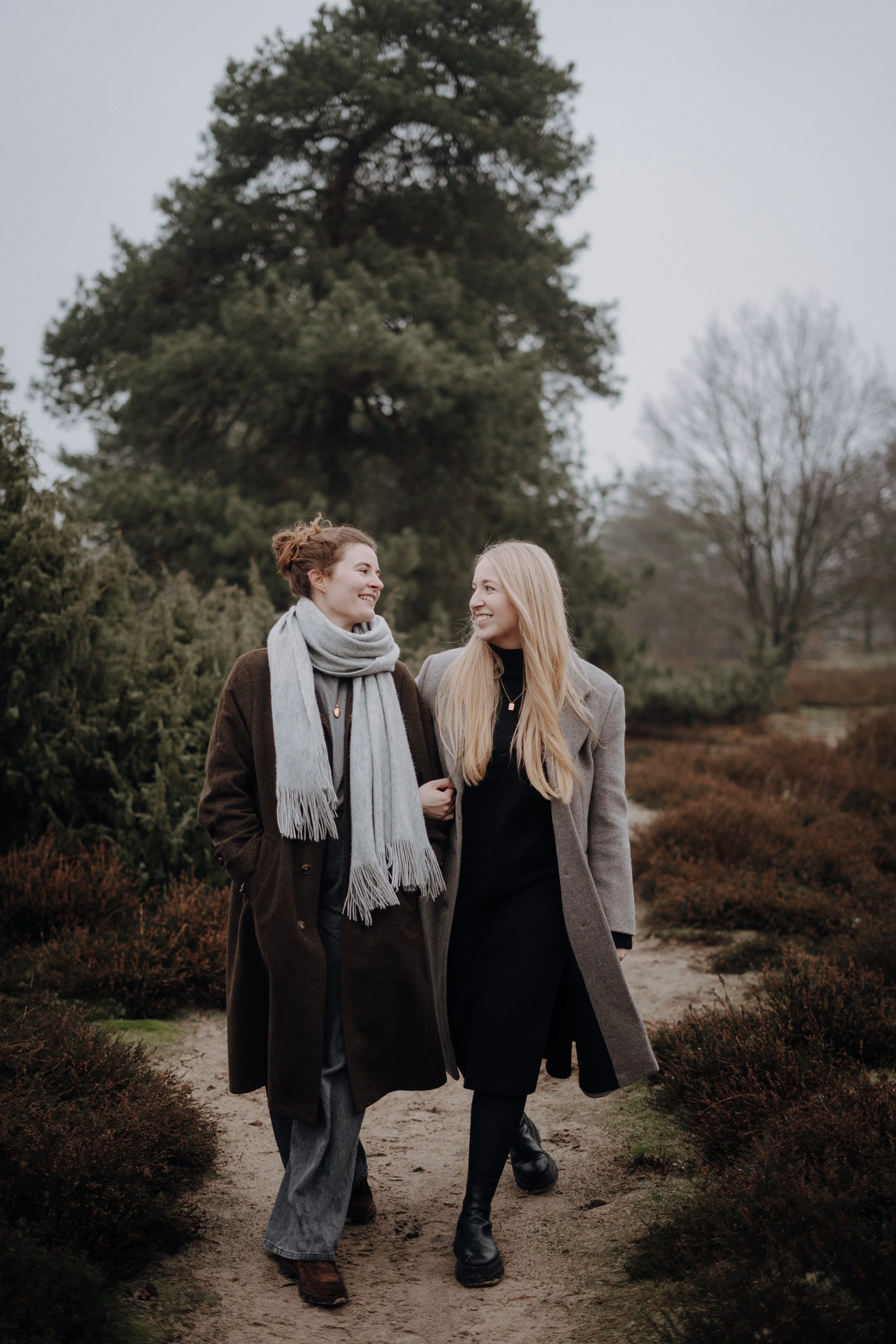 Zwei Frauen spazieren an einem bewölkten Tag durch eine parkähnliche Umgebung mit Büschen und Bäumen, lächelnd und sich gegenseitig anlächelnd.