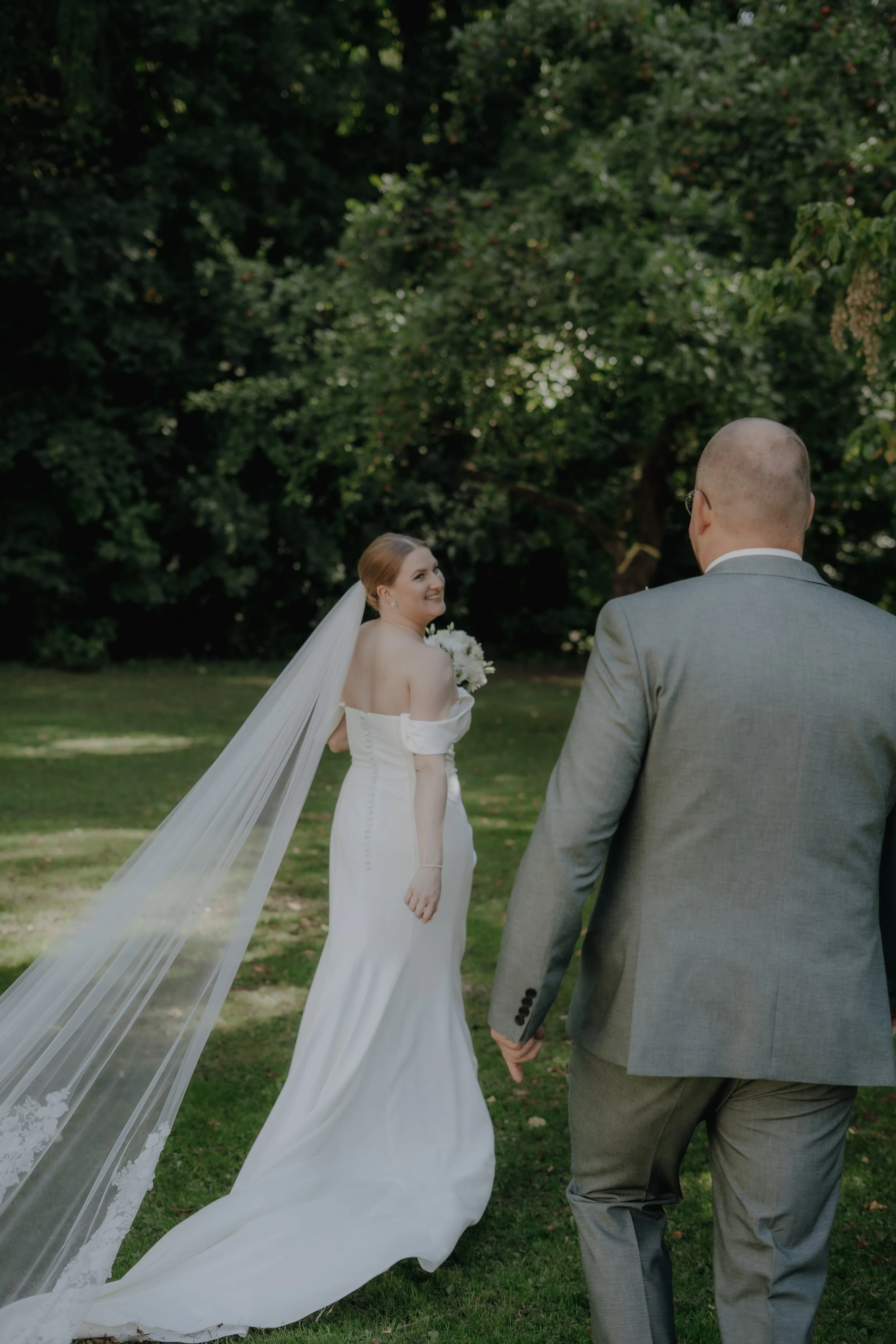 Braut und Bräutigam im Park, Blick auf die Braut, die in einem weißen Brautkleid mit langen Schleppe und Schleier steht. Sie lächelt und schaut zum Bräutigam, der in einem grauen Anzug steht.