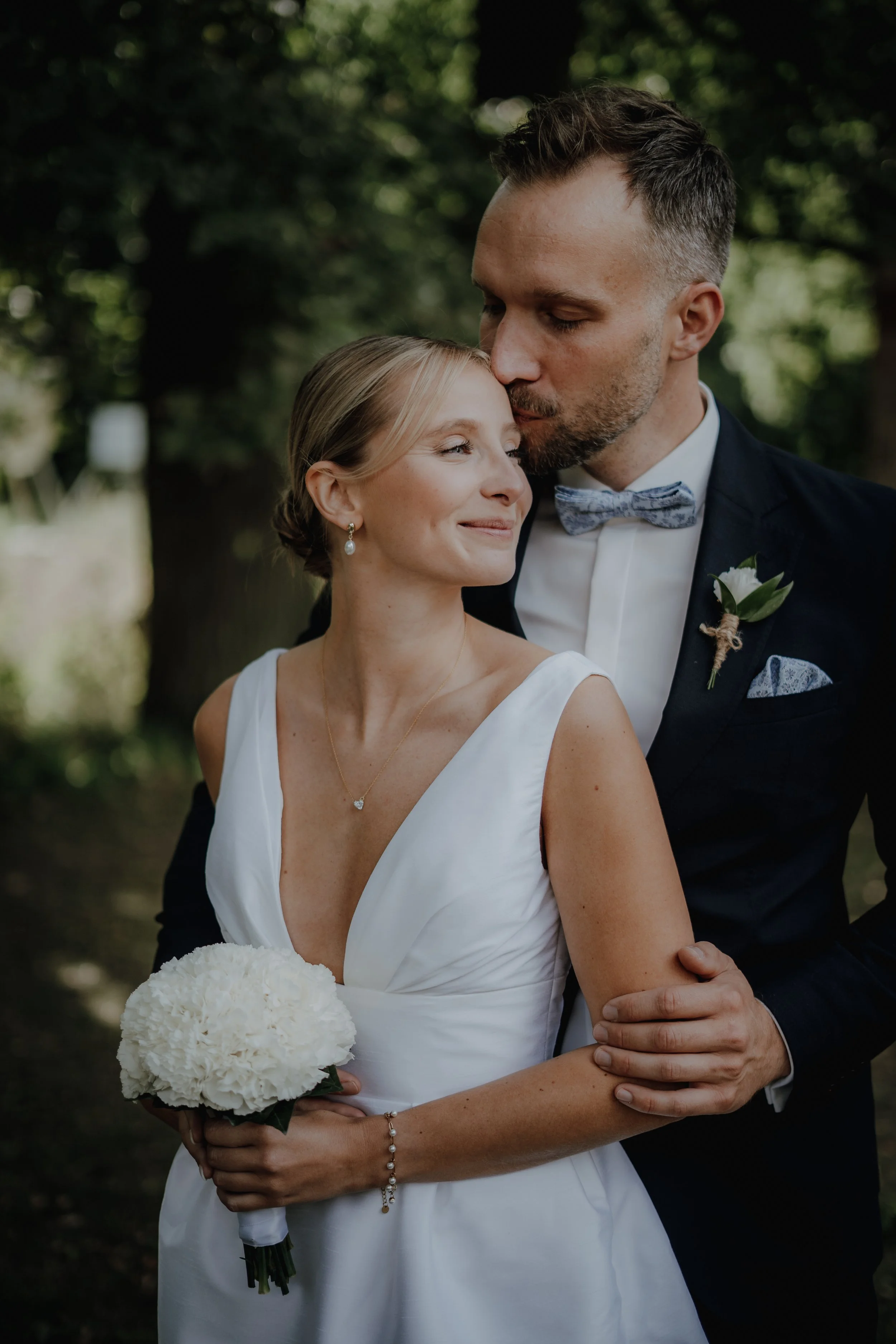 Ein Brautpaar bei einer Hochzeit im Freien. Die Braut trägt ein weißes Kleid und hält einen Blumenstrauß, der Bräutigam trägt einen dunklen Anzug mit Fliege. Der Mann küsst die Braut auf die Stirn.