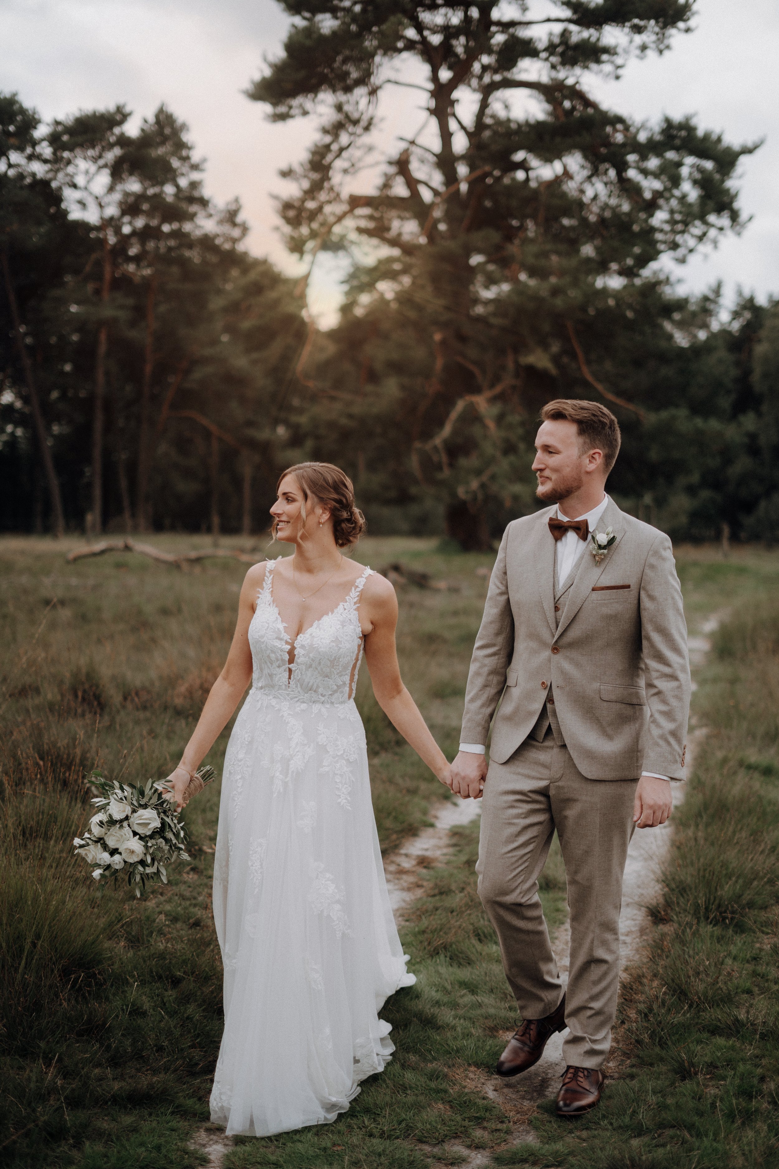 Verliebtes Brautpaar hält Händchen auf einem Waldweg bei Sonnenuntergang.