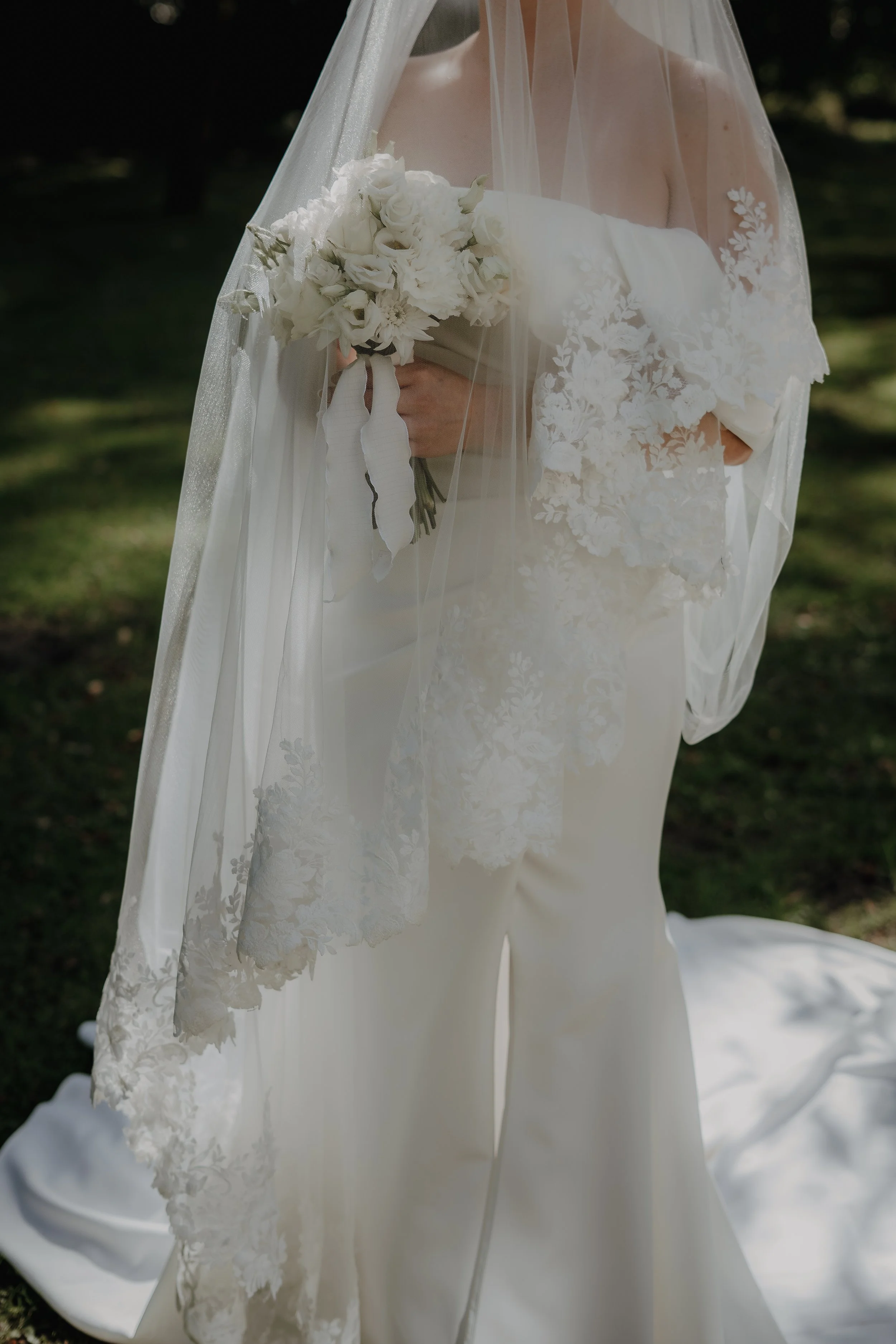 Braut in weißem Hochzeitskleid und Schleier hält einen Brautstrauß aus weißen Blumen im Freien.