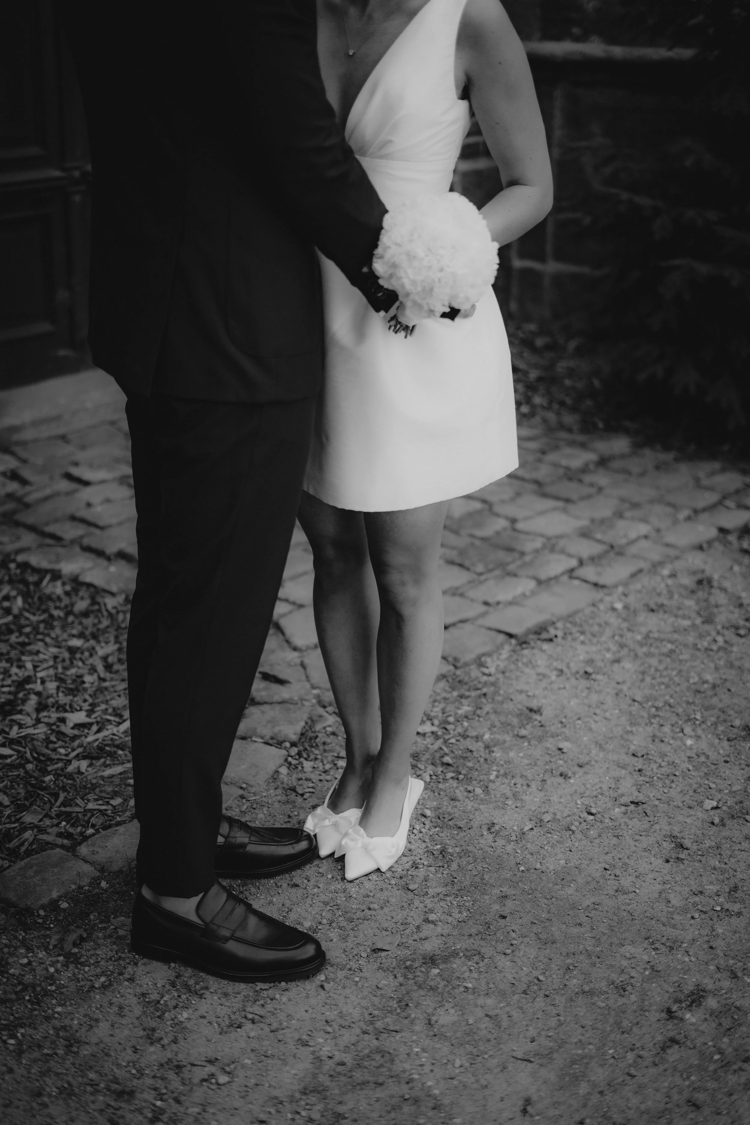 Ein Brautpaar bei einer Hochzeit, Nahaufnahme der unteren Körperhälfte, schwarz-weiß, auf Cobblestone und Erdweg stehend.