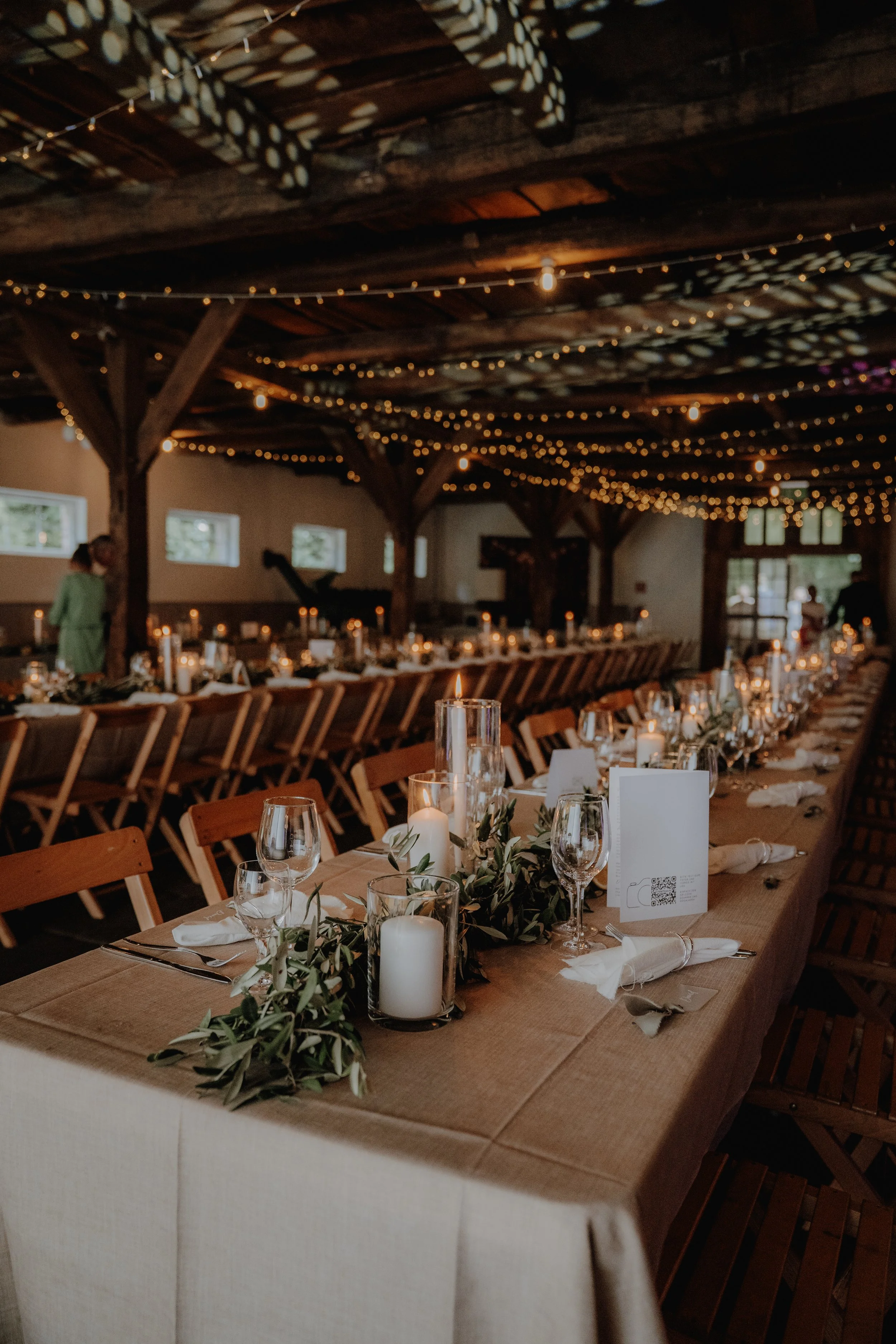 Ein festlich gedeckter langer Tisch in einer rustikalen Scheune mit Kerzen, Grünzeug und weihnachtlicher Beleuchtung für eine Feier.