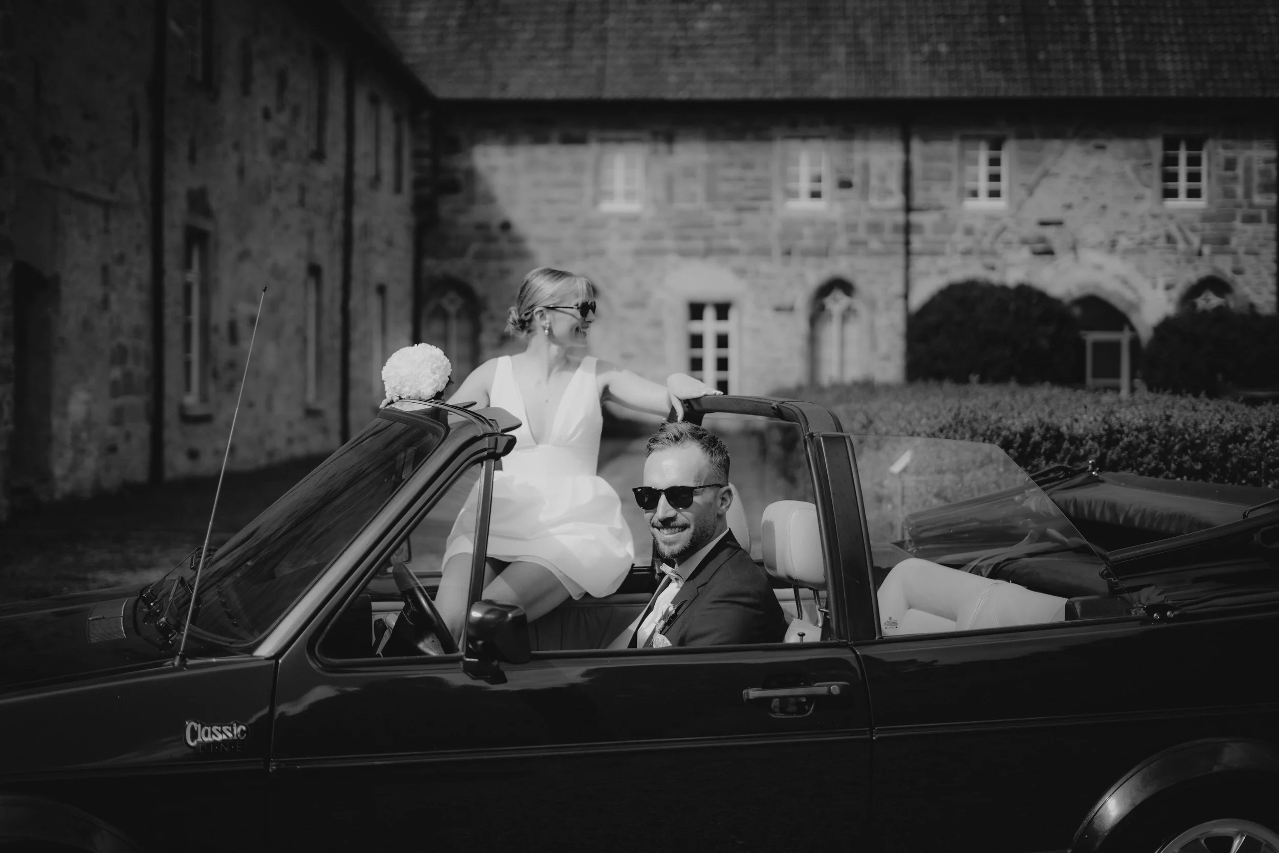 Ein Mann und eine Frau fahren in einem Cabrio. Der Mann trägt einen Anzug und Sonnenbrille, die Frau trägt ein weißes Kleid und Sonnenbrille. Es scheint eine Hochzeit zu sein.