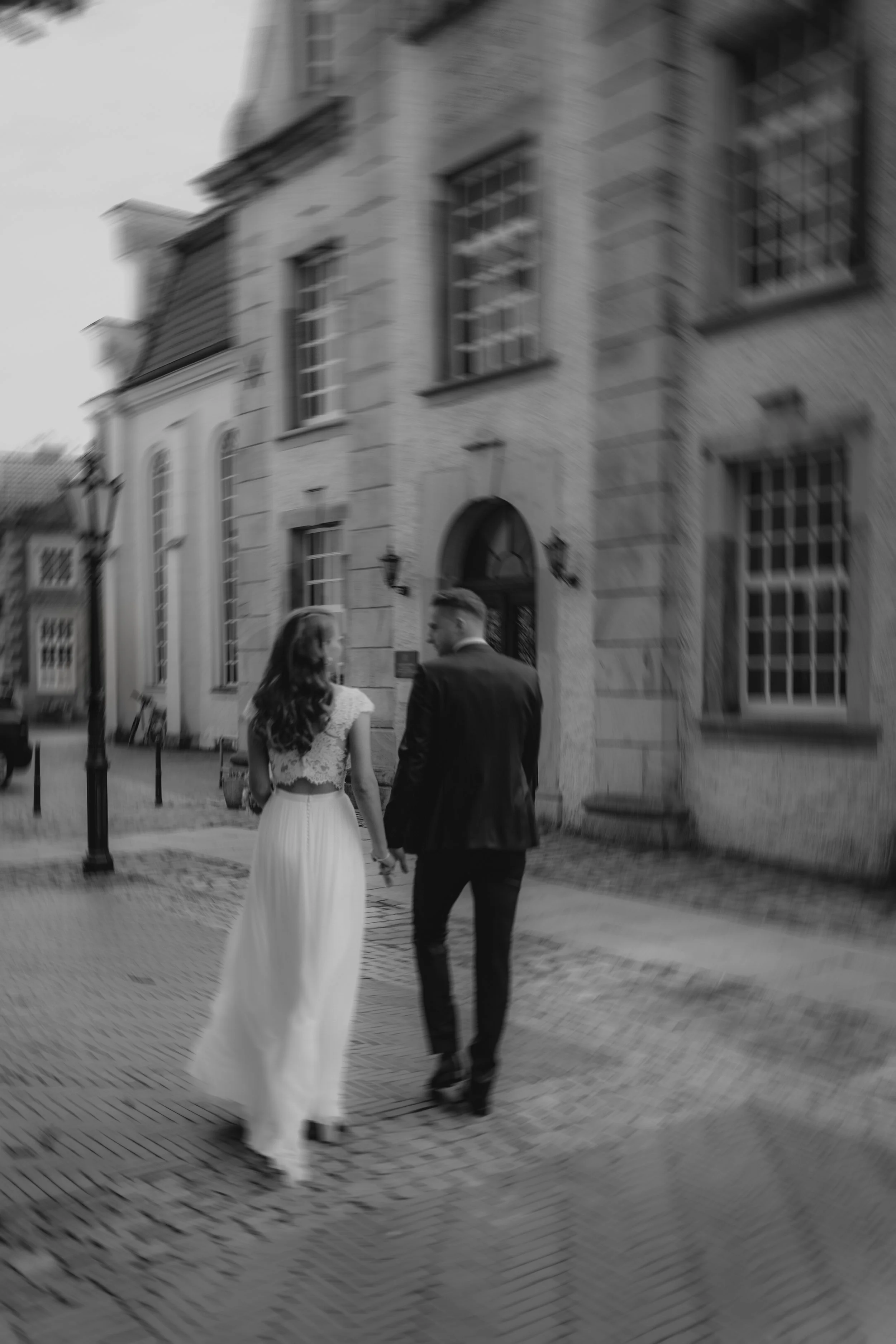 Ein verliebtes Paar läuft Hand in Hand vor einem historischen Gebäude in Schwarz-Weiß, wahrscheinlich bei einer Hochzeit oder einem besonderen Anlass.