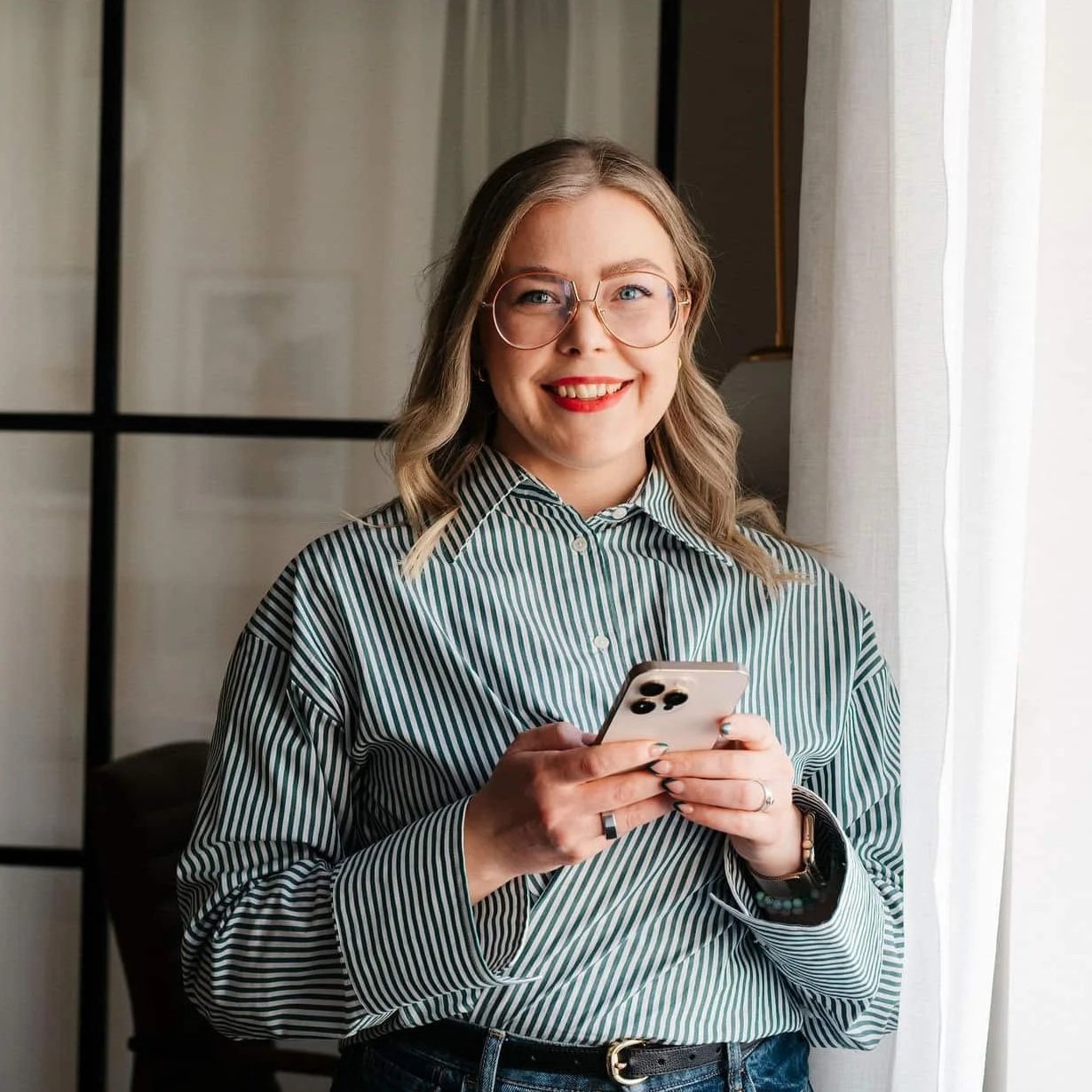 Nainen pitää älypuhelinta ja hymyilee ikkunan äärellä.