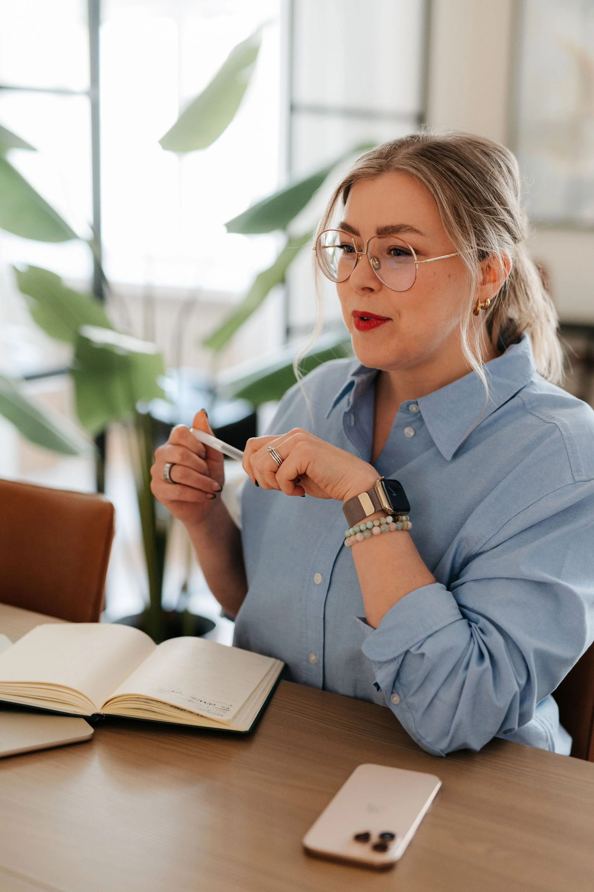 Nainen istuu pöydän ääressä avointa muistikirjaa katsellen, housupaita päällä, kello ja koruja ranteessa, suurilla silmälaseilla, ja taustalla kasvi ikkunan läpi.