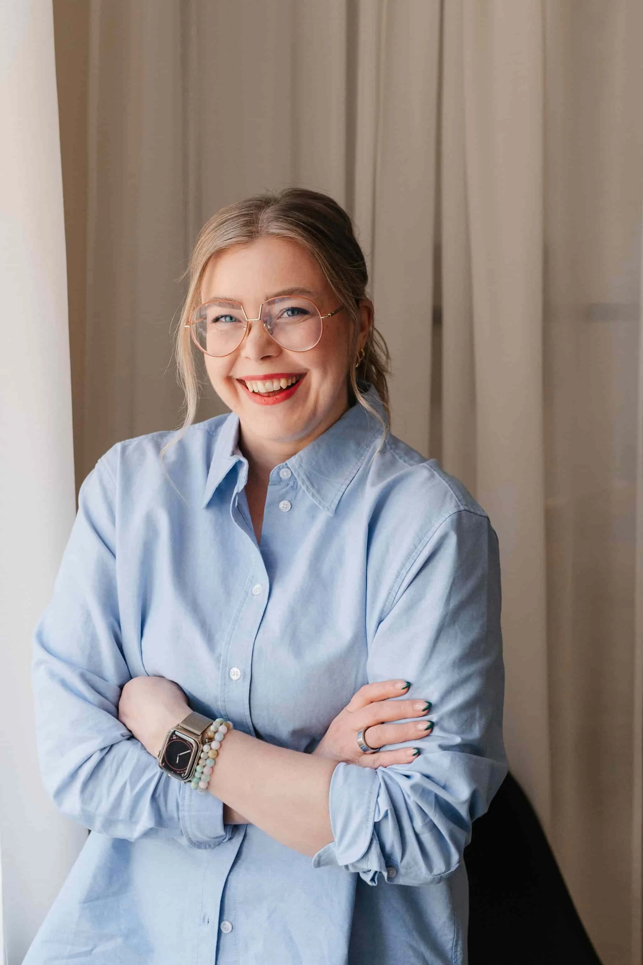 Nuorehko nainen hymyilee, istuu ikkunan vieressä ja pitää käsivarsiensa ristiin.