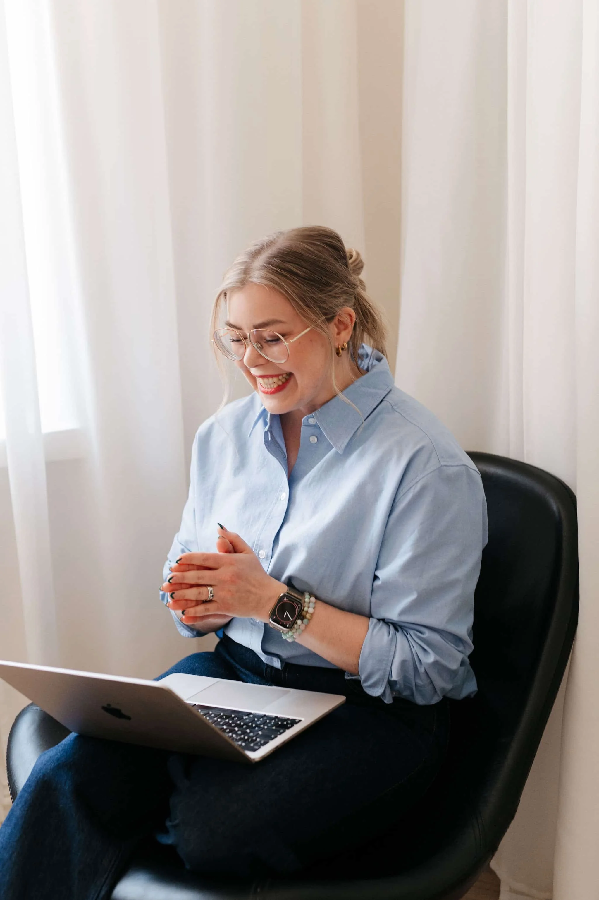 Nainen istuu tuolilla käyttäen avoimena kannettavaa tietokonetta ja hymyilee.