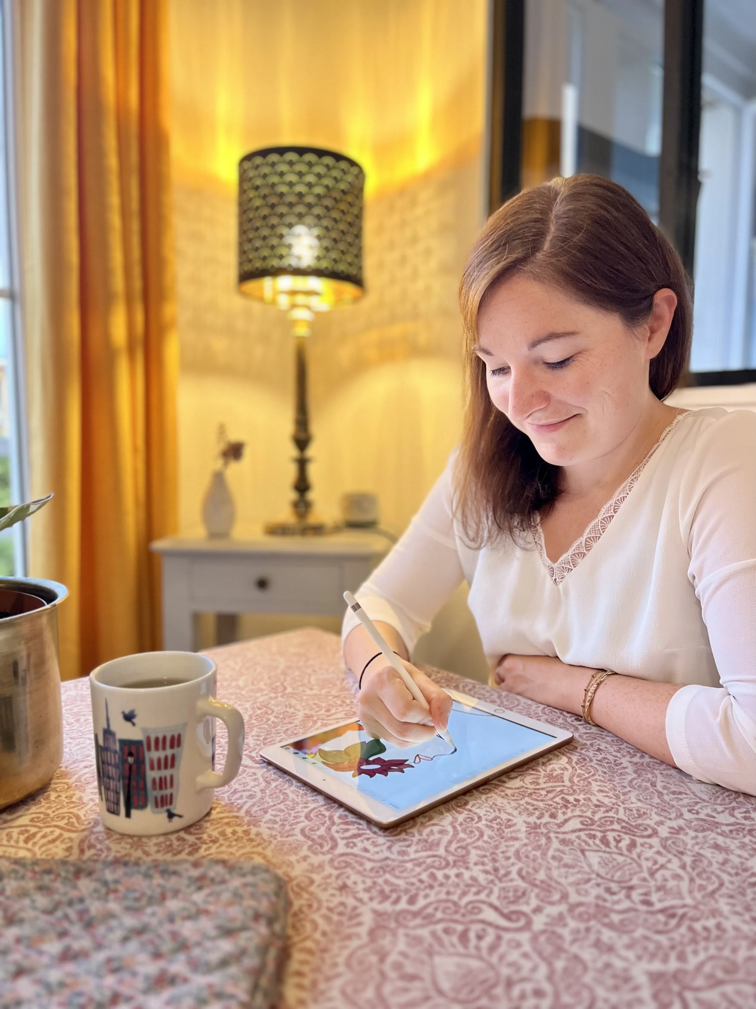 Une femme dessinant sur une tablette numérique à une table avec une tasse, une plante en métal, et un décor intérieur chaleureux avec lampe et rideaux jaunes.