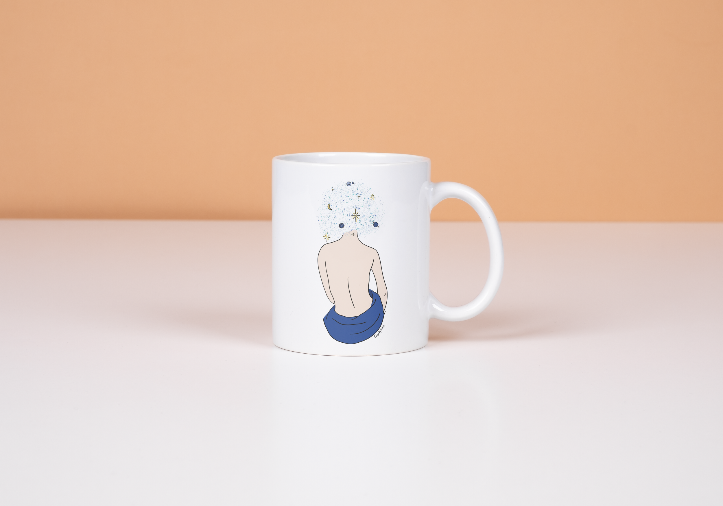 Une tasse blanche avec une illustration d'un dos nu d'une personne avec un drapé bleu, dont la tête est remplacée par un motif cosmique composé d'étoiles, de planètes et de la lune, sur fond beige clair.