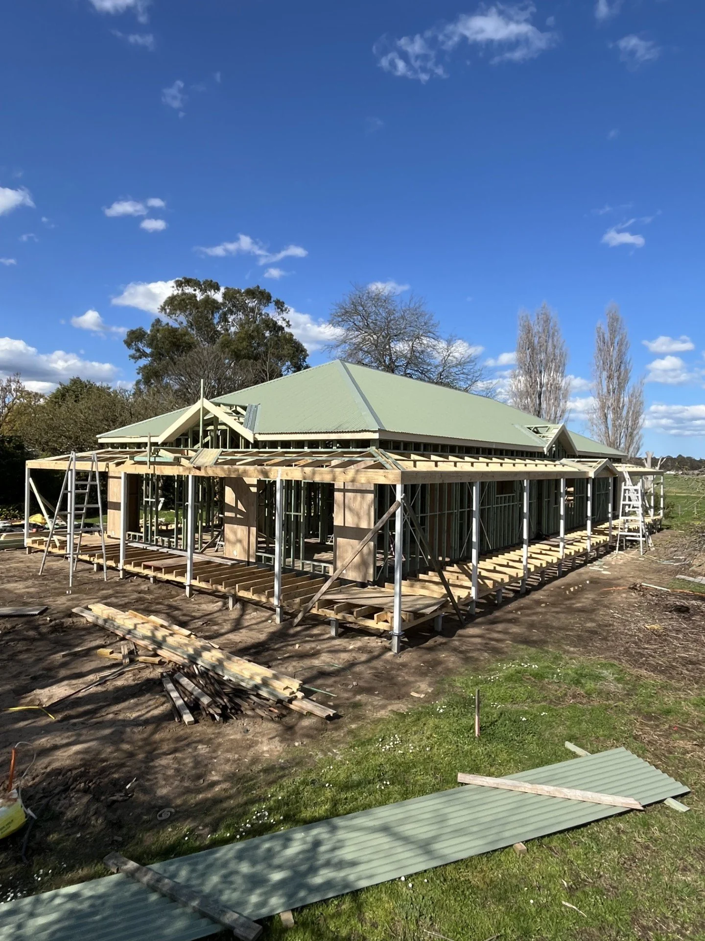 Construction site of a house with a green roof, wooden framework, and scaffolding, surrounded by trees and blue sky.