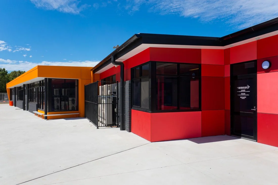 Colorful school exterior with orange and red panels, large windows, black framing, and a clear blue sky.