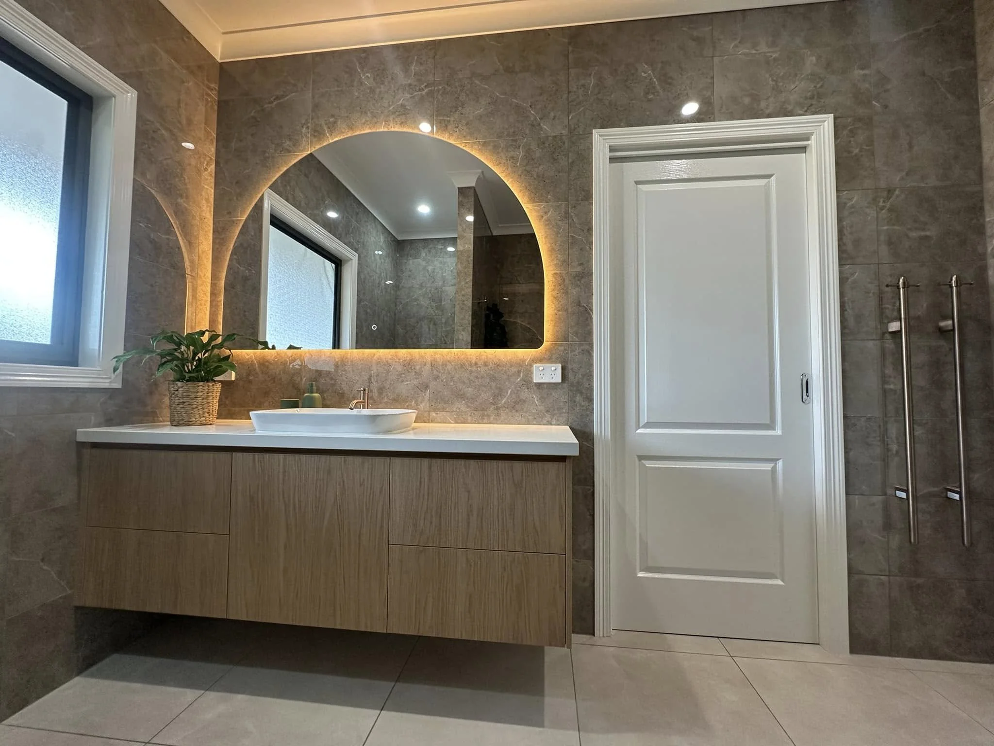 A modern bathroom with a large backlit mirror, a wooden vanity with a white sink, a potted plant, frosted windows, and a closed white door.