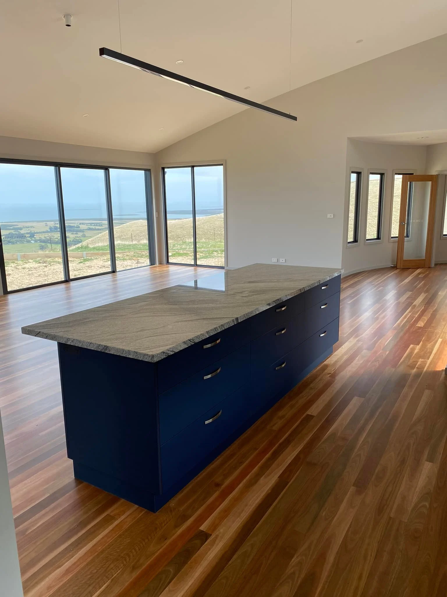 Modern open-concept living area with large glass sliding doors, a kitchen island with a marble top, dark blue cabinetry, hardwood floors, and large windows showing an outdoor landscape with hills and water.
