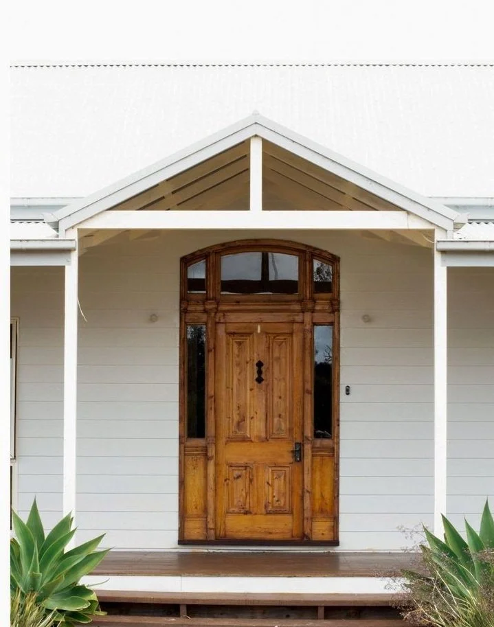 Front view of a house with a large wooden front door, white siding, a small front porch, and green plants on either side.