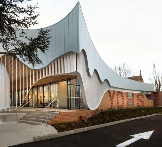 Unique modern building with wavy exterior, large glass windows, steps leading to entrance, and a landscaped area in front.