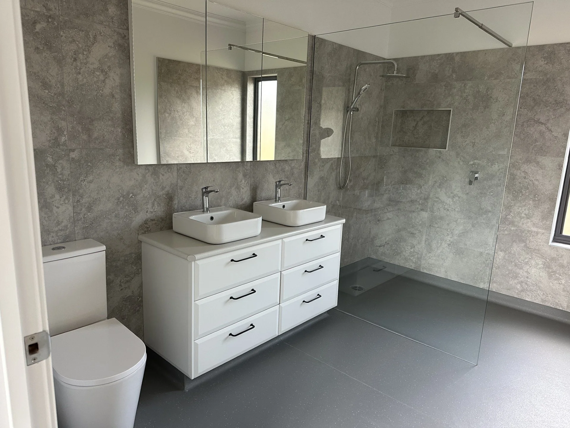 Modern bathroom with gray tiled walls, a double vanity with two square sinks, a large mirror, a toilet, and a glass-enclosed walk-in shower with a rainfall showerhead and a window.