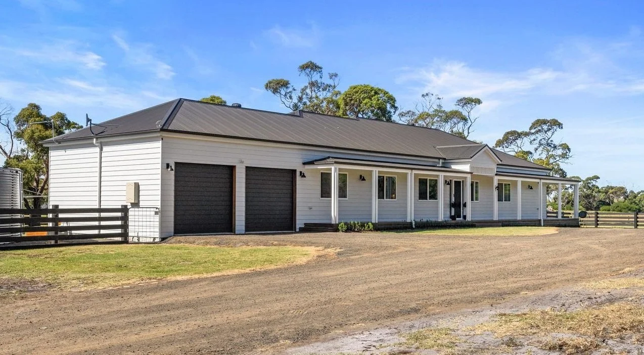 Modern country style house with white siding, black garage doors, and a grey metal roof, set against a background of blue sky and trees.