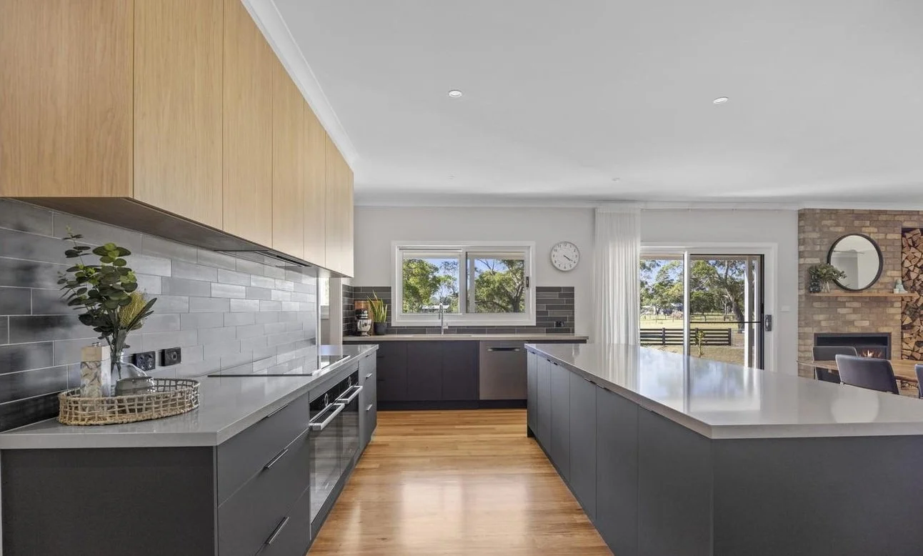 Modern kitchen with gray cabinets, a large island, wood flooring, and a brick fireplace in the background.