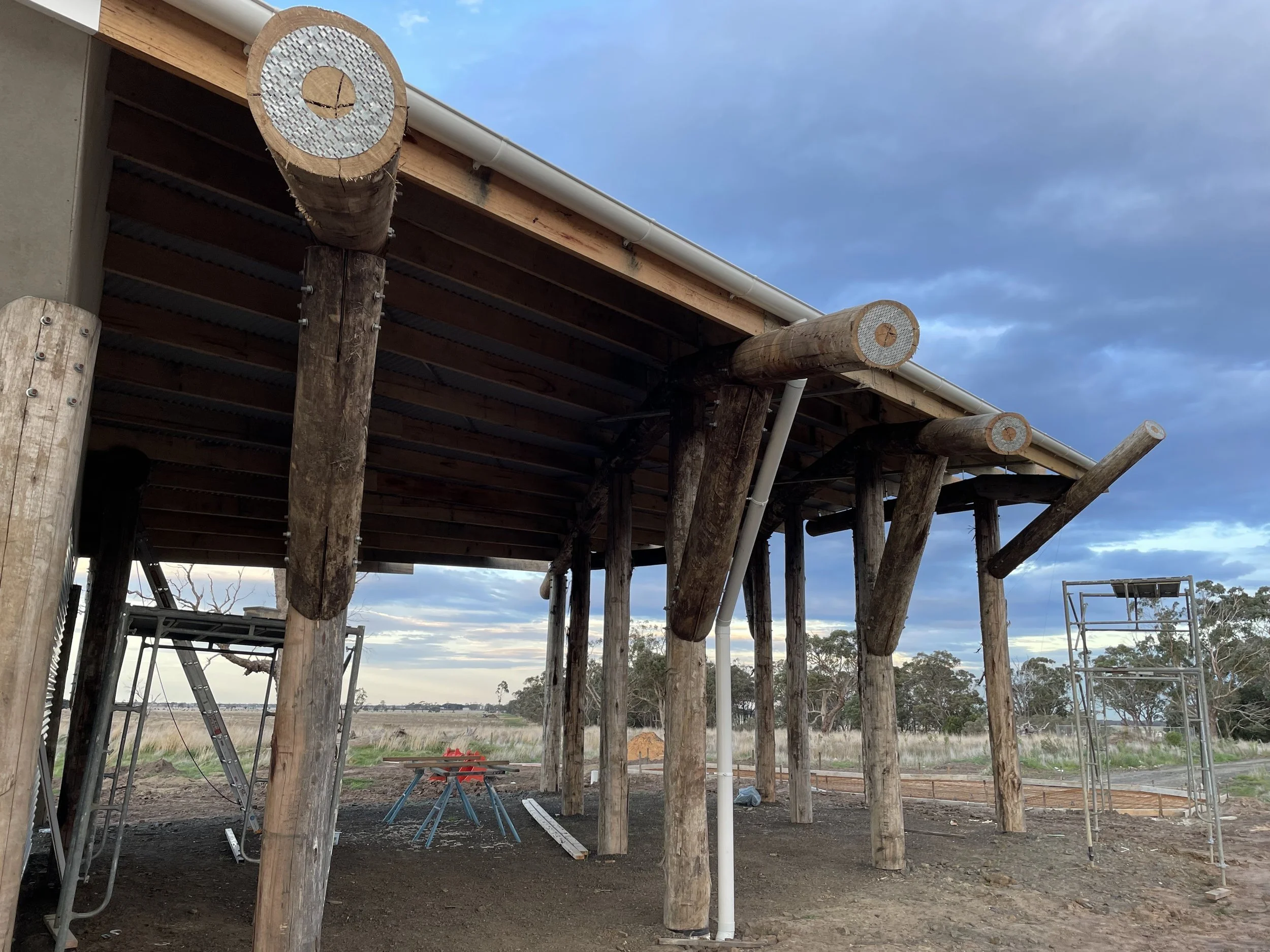 Construction site with wooden support beams and scaffolding under a raised structure, against a partly cloudy sky.