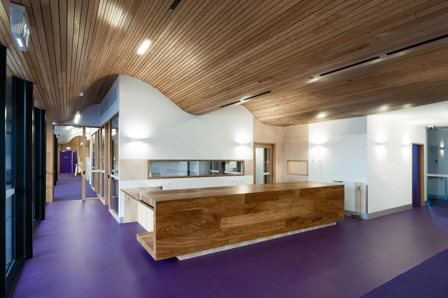 Modern interior space with curved wooden ceiling, white walls, purple flooring, and a wooden reception desk.