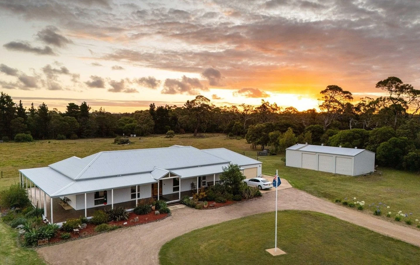 A white house with a metal roof, a gravel driveway, a flagpole with an Australian flag, and a large rural yard at sunset or sunrise.