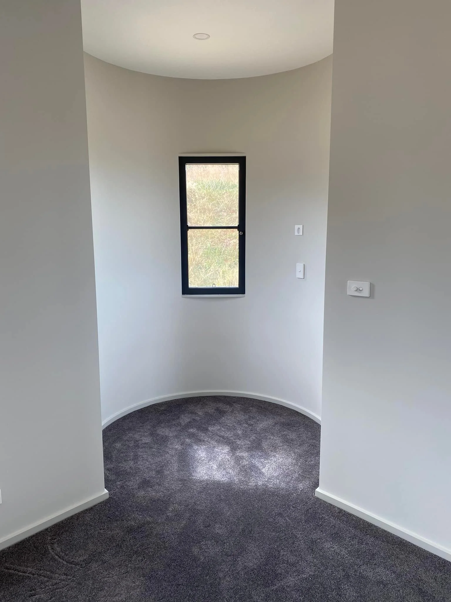 Room with small window, white walls, gray carpeting, and three electrical switches on the wall.