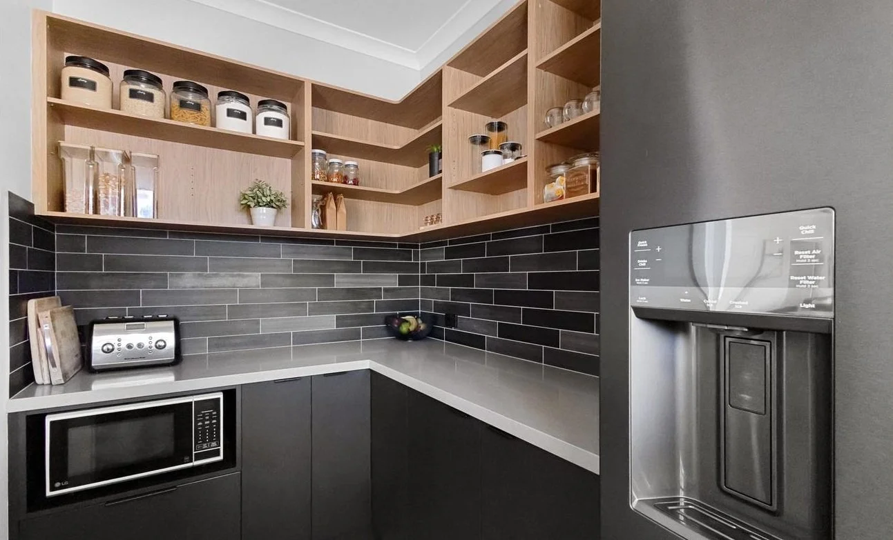 Modern kitchen corner with black and gray cabinets, black tiled backsplash, open wooden shelves with jars and decorative items, microwave, toaster, and a bowl of apples.