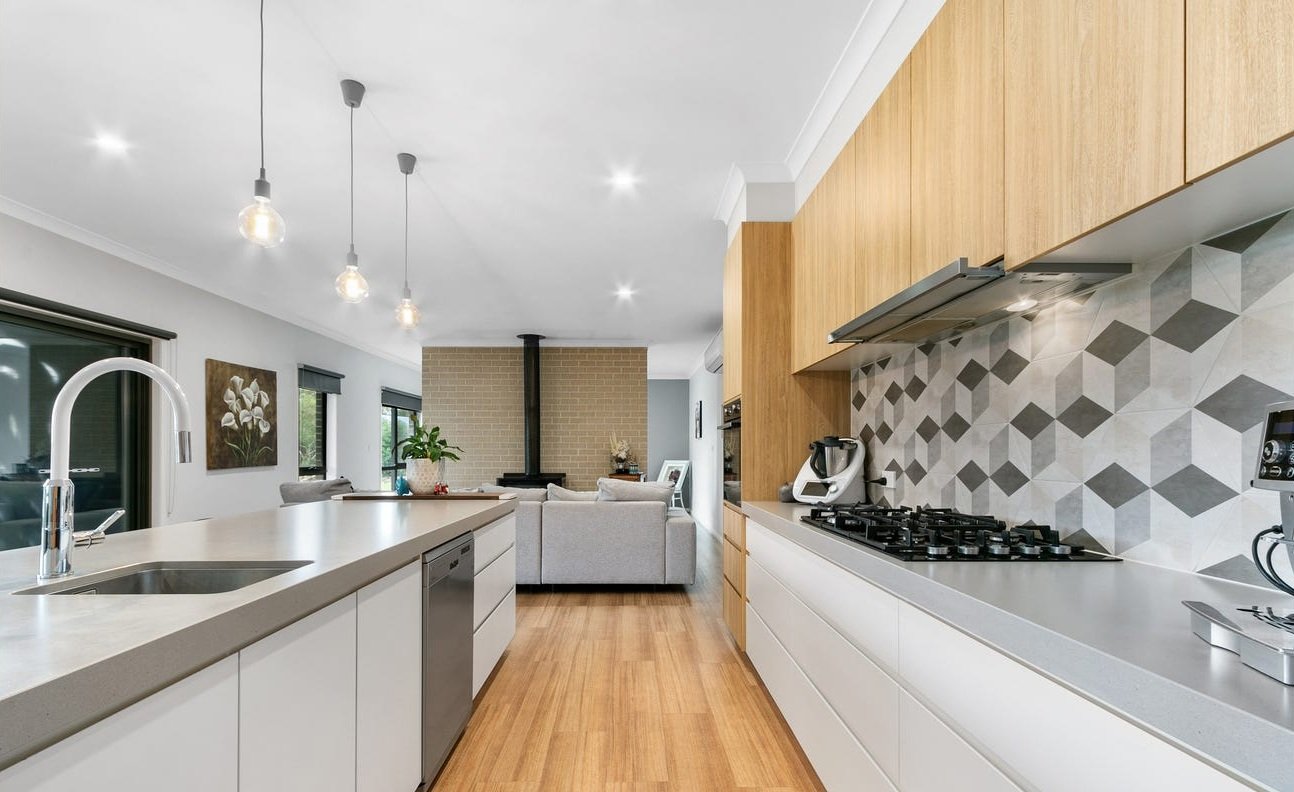 Modern kitchen with white countertops, wooden cabinetry, and a geometric gray tile backsplash. The kitchen features a stainless steel sink, built-in appliances, and pendant lights hanging from the ceiling. The open layout reveals a living room with g