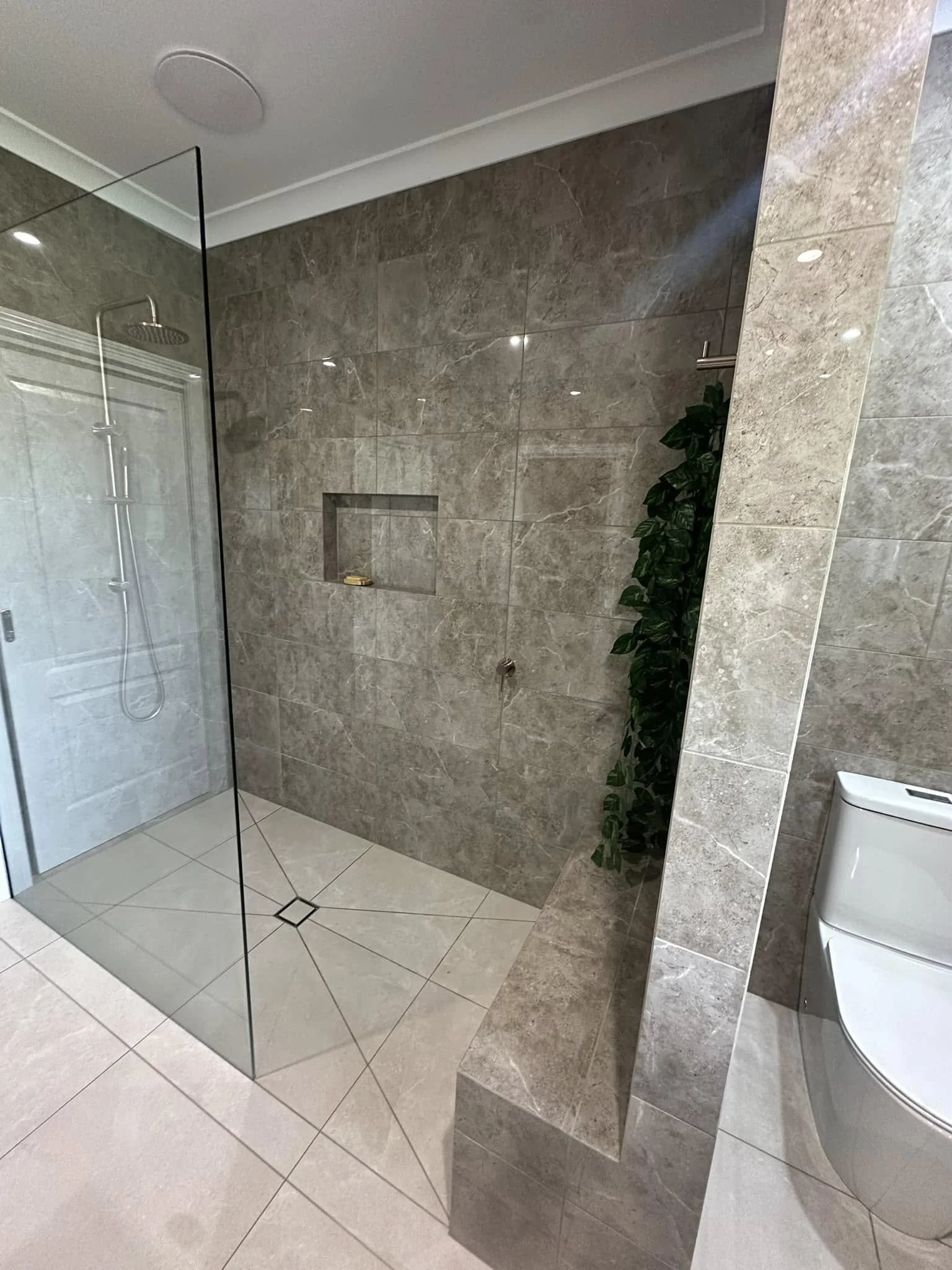 Modern bathroom with a walk-in shower enclosed by glass, a built-in shower niche, a white toilet, and a tall green plant, all with beige and gray tile surroundings.