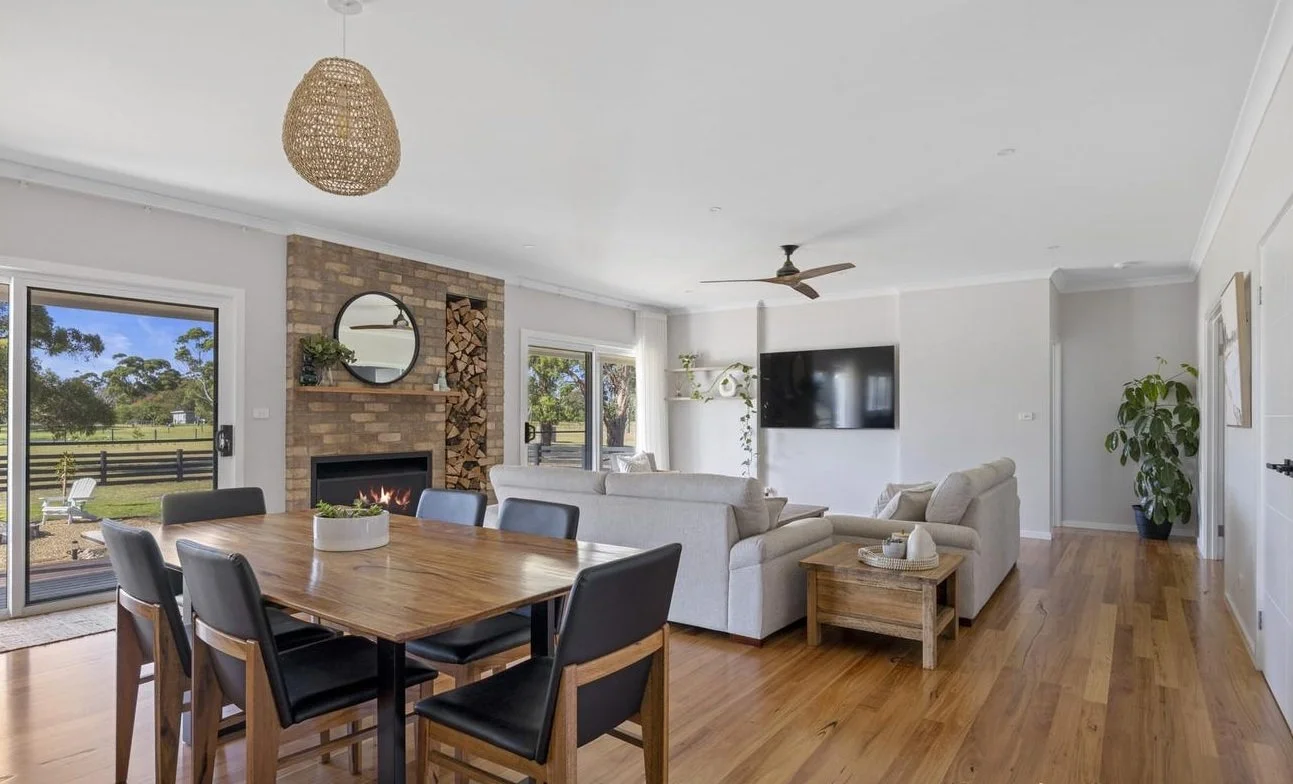 Open living and dining room with hardwood floors, white walls, a brick fireplace, a sliding glass door to the outside, a mounted flat-screen TV, and a ceiling fan.