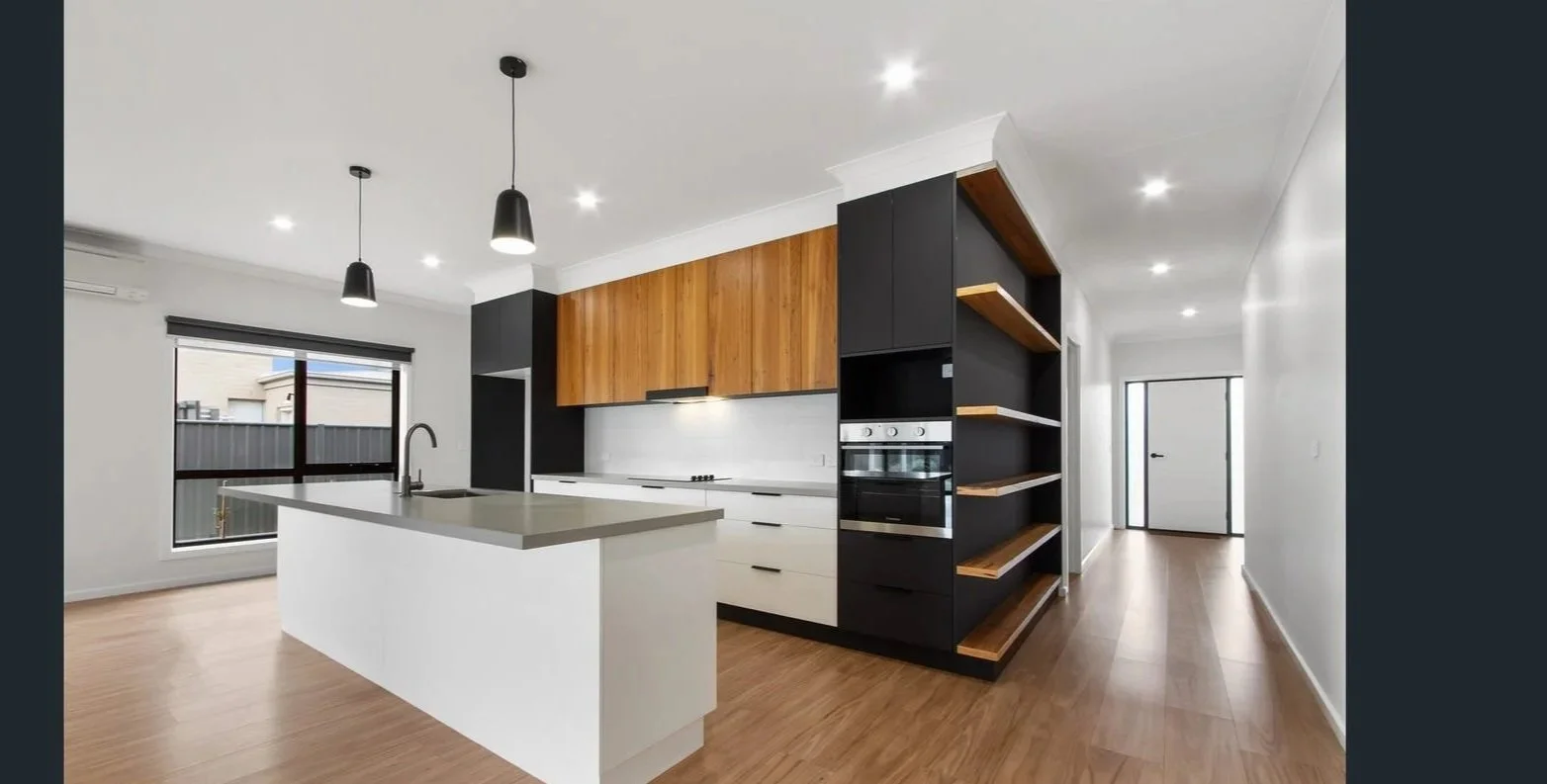 Modern kitchen with white island, black and wooden cabinets, built-in oven, large window, and wooden flooring.