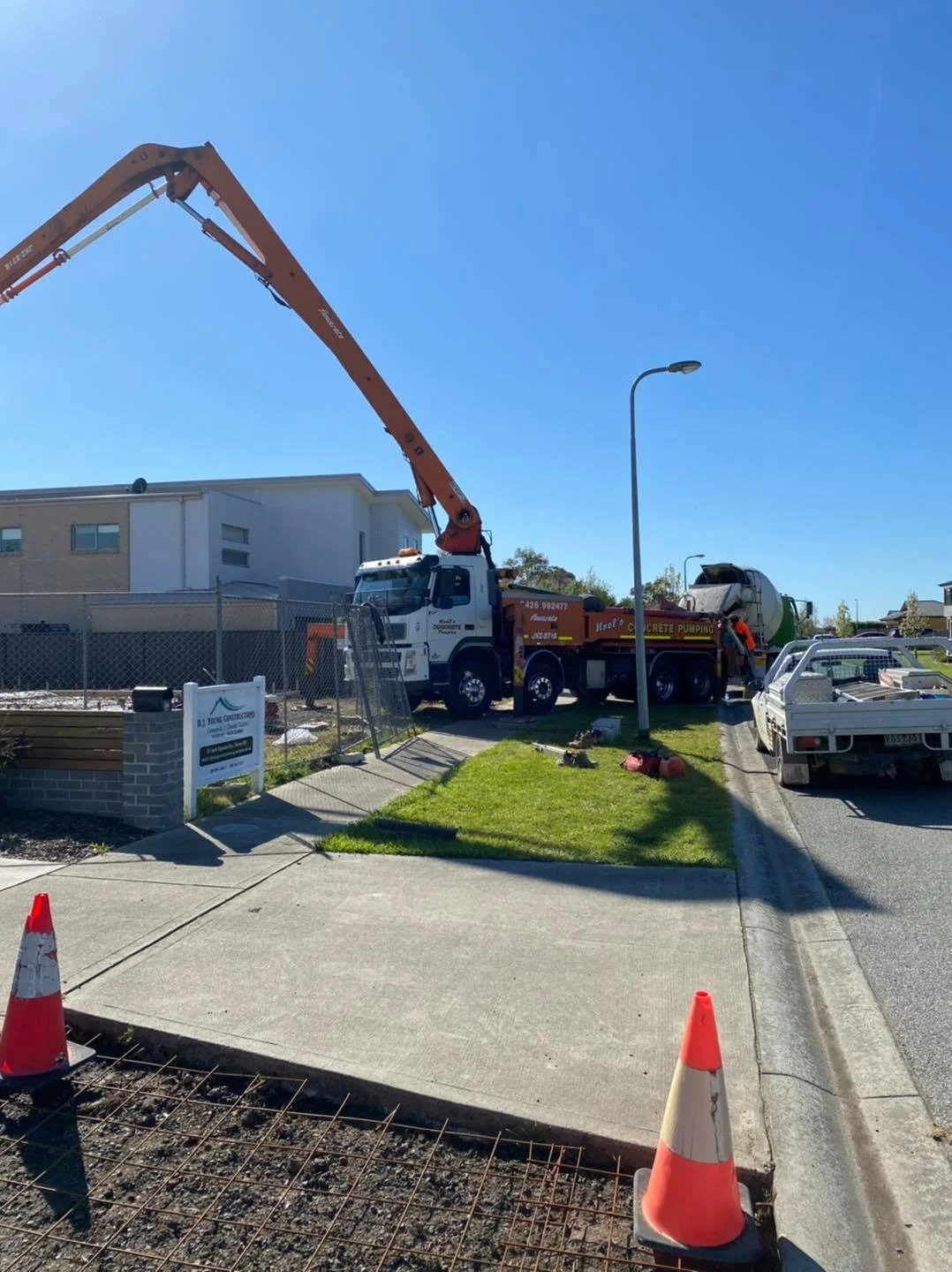 Construction site with a concrete pump truck, orange and white safety cones, a cement mixer truck, and a pickup truck, near a modern residential building on a sunny day.